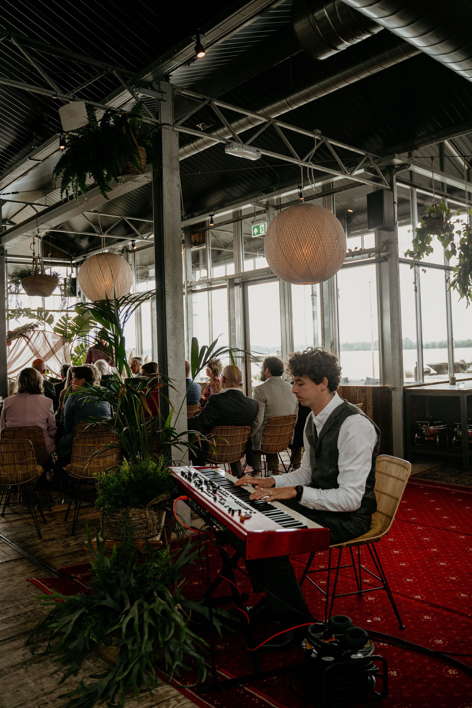 A musician performs on a keyboard in an industrial-style venue with paper lanterns and plants.