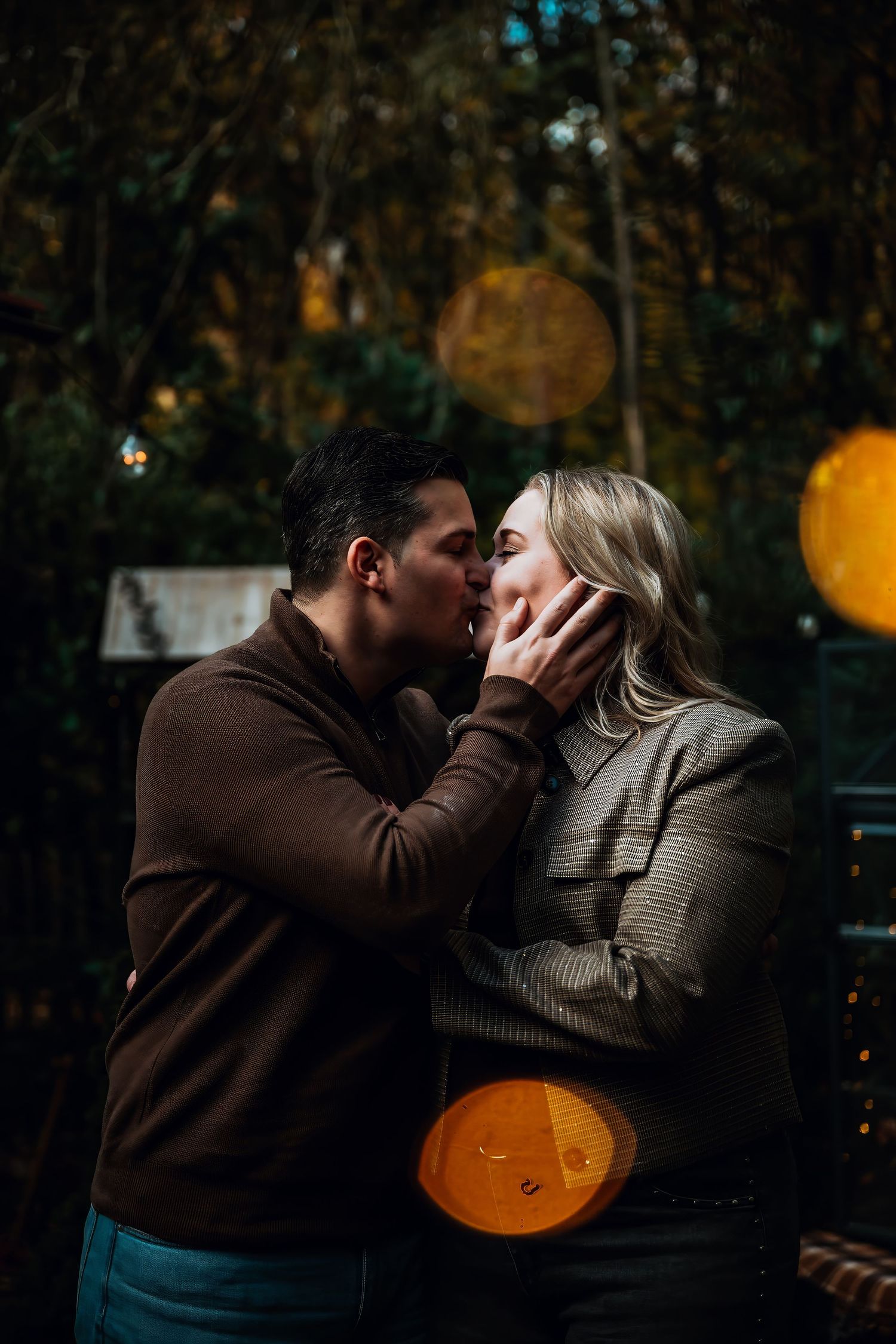 A romantic couple embraces in a dark outdoor setting with glowing orange bokeh lights in the background.