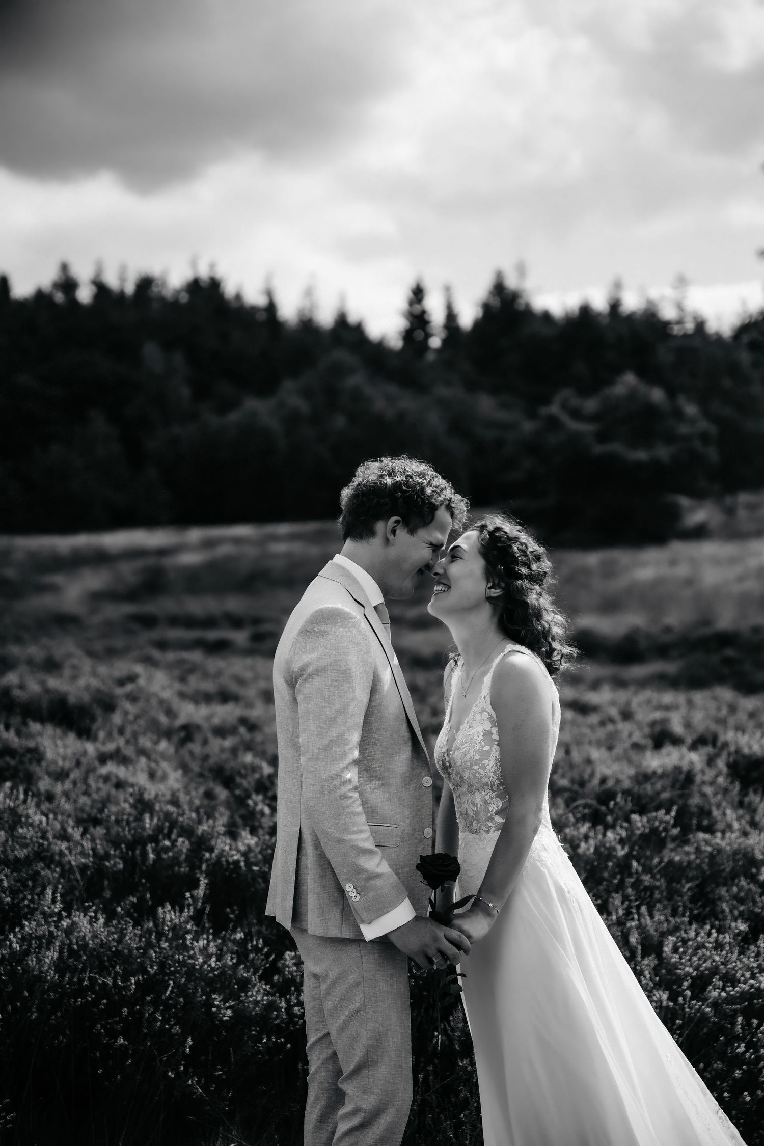 Romantic black and white portrait of a couple embracing in a field with dramatic cloudy sky.