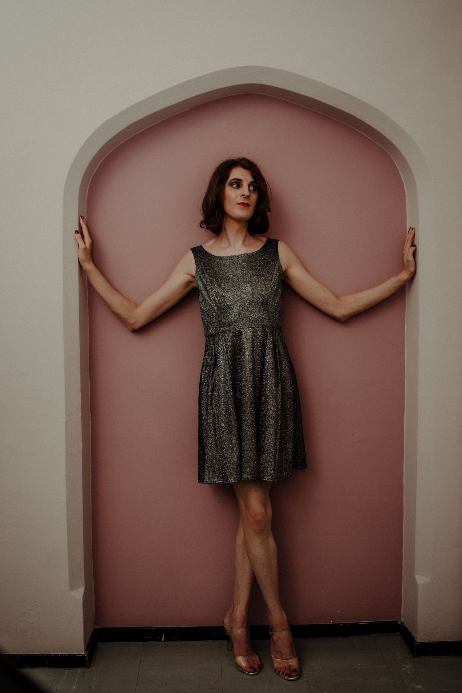 A person in a sparkly dress stands against a pink arched alcove in various dramatic poses.