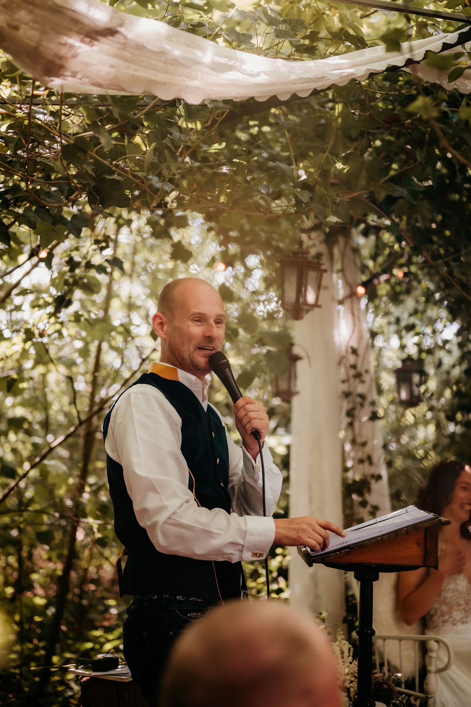 A speaker in formal attire delivers remarks at an outdoor ceremony under a canopy of trees and natural light.