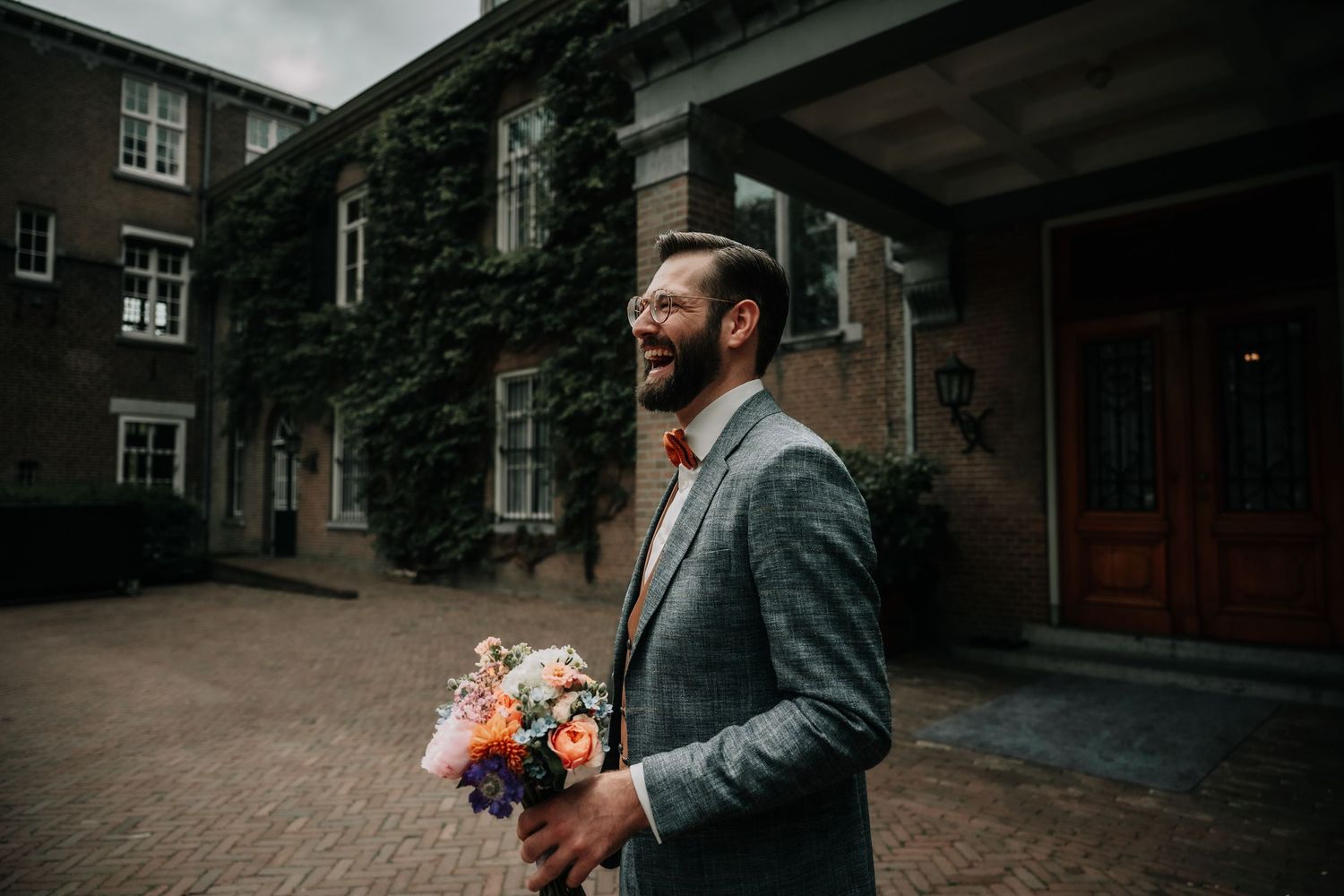 Person in grey suit jacket holding colorful bouquet while smiling outside brick building covered in ivy.