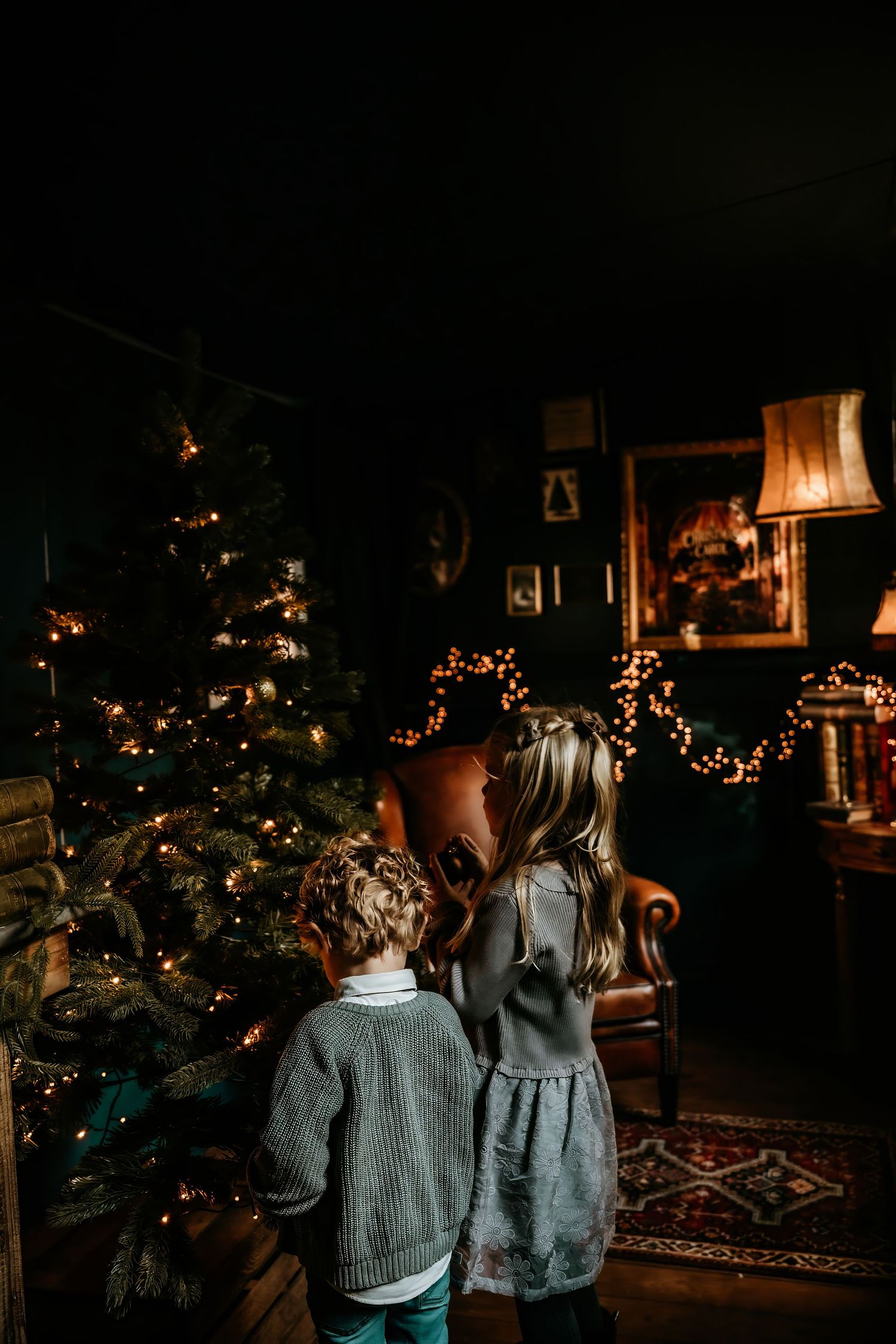 Children in matching sweaters admire a glowing Christmas tree in a dark room at night.
