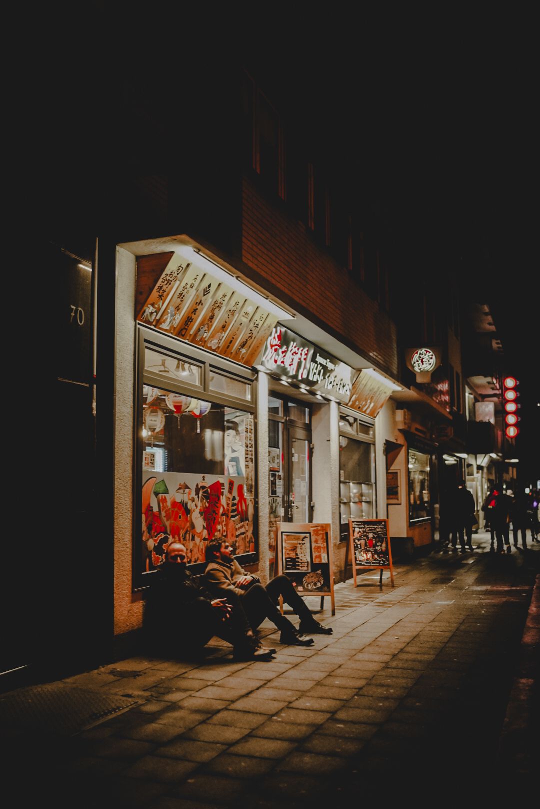 Dimly lit street at night with illuminated storefronts and red lanterns casting a warm glow on the sidewalk.