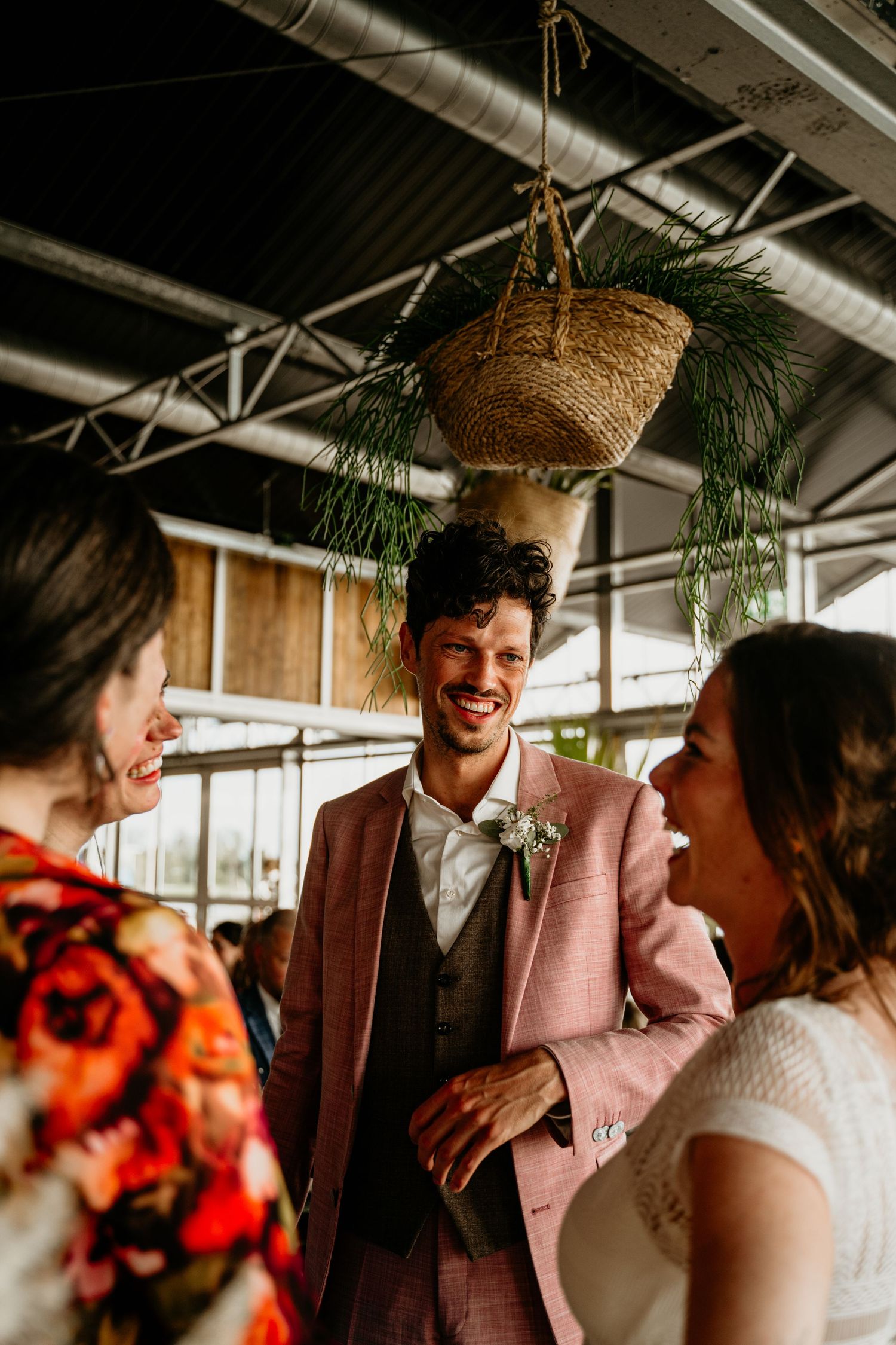 Wedding guests share laughter under hanging greenery and woven basket decor in an industrial space.