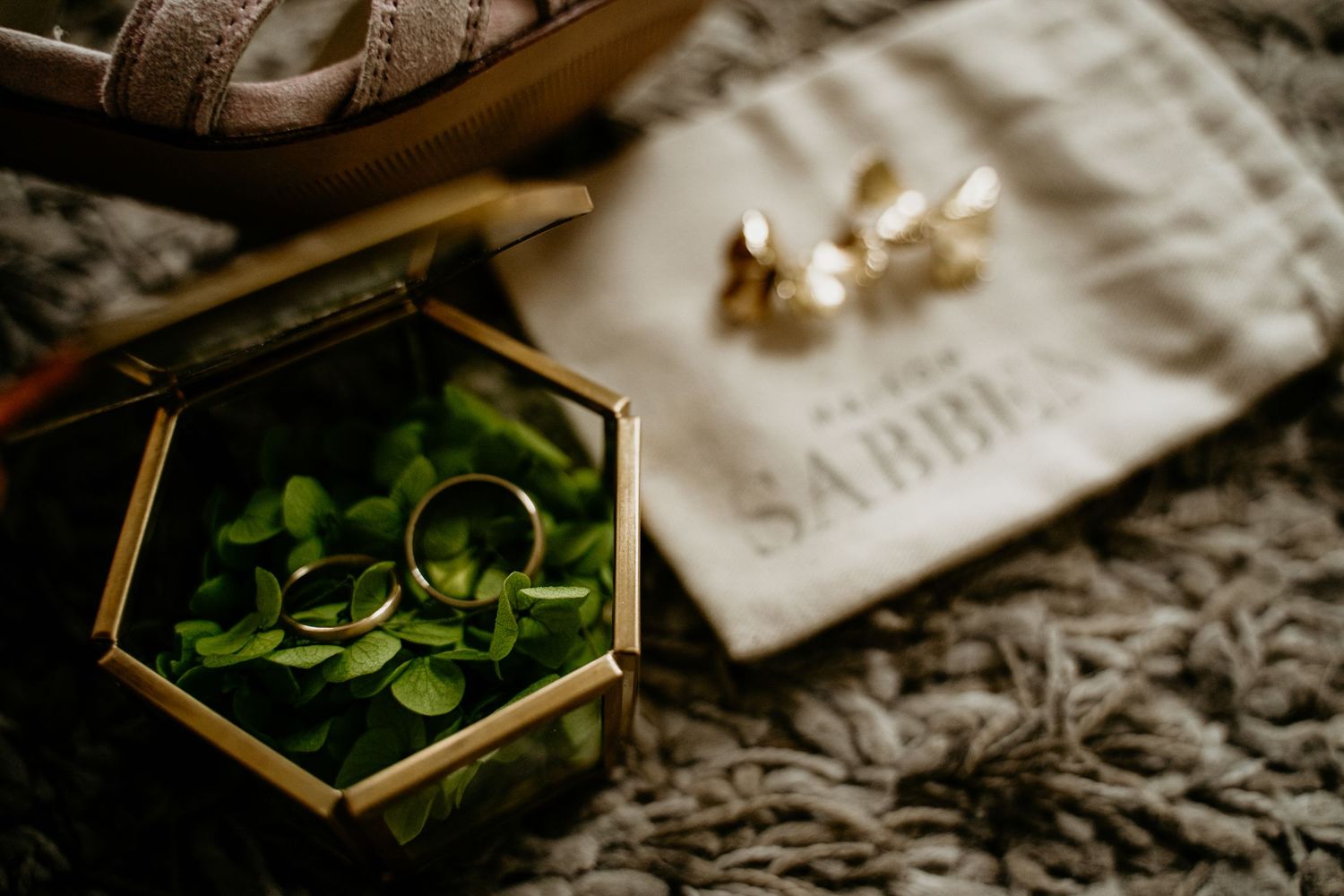 Wedding rings rest beside a gold hexagonal box filled with fresh green leaves on a textured fabric surface.
