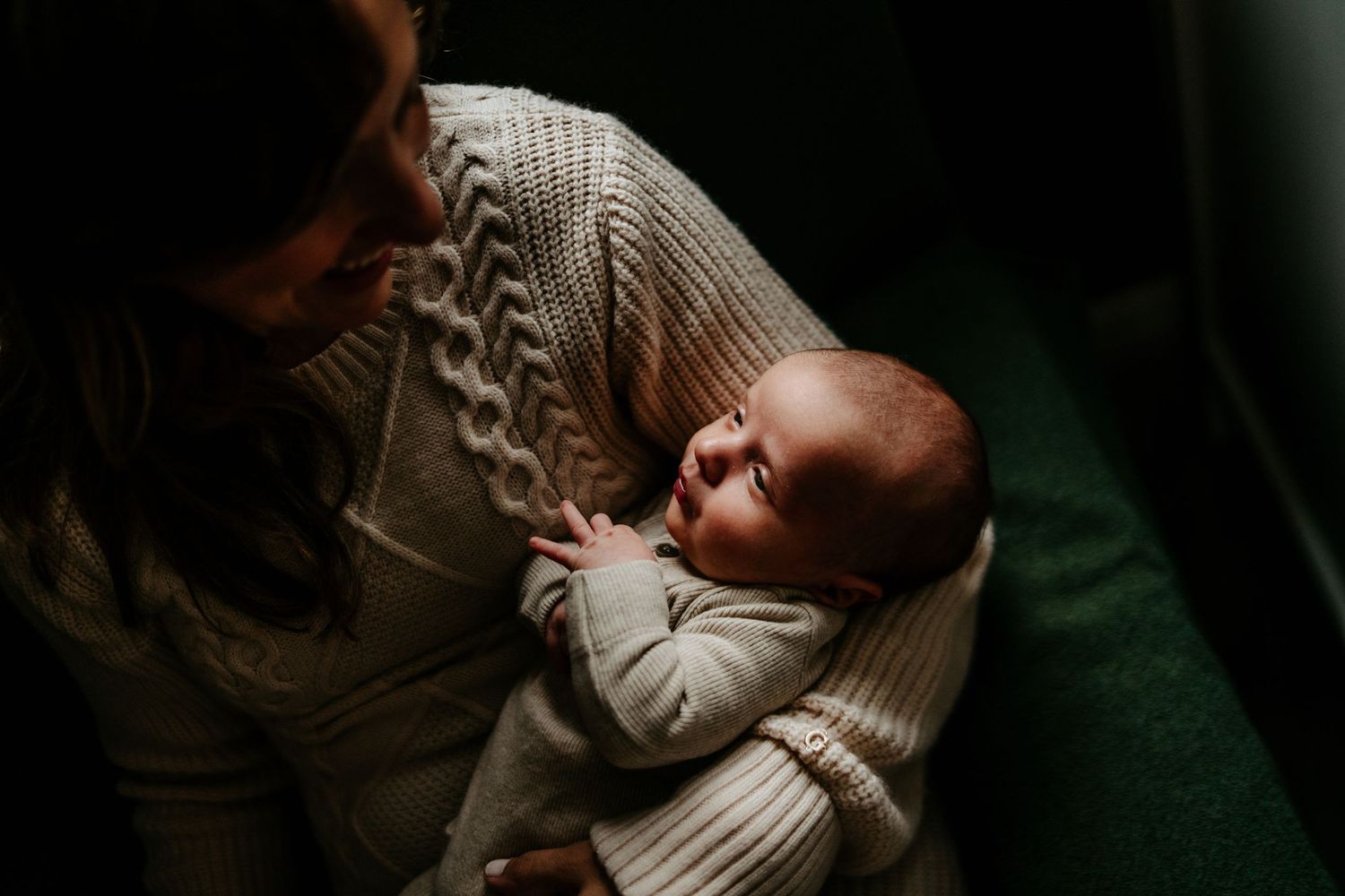 Mother in cable knit sweater holding sleeping newborn in dim lighting.