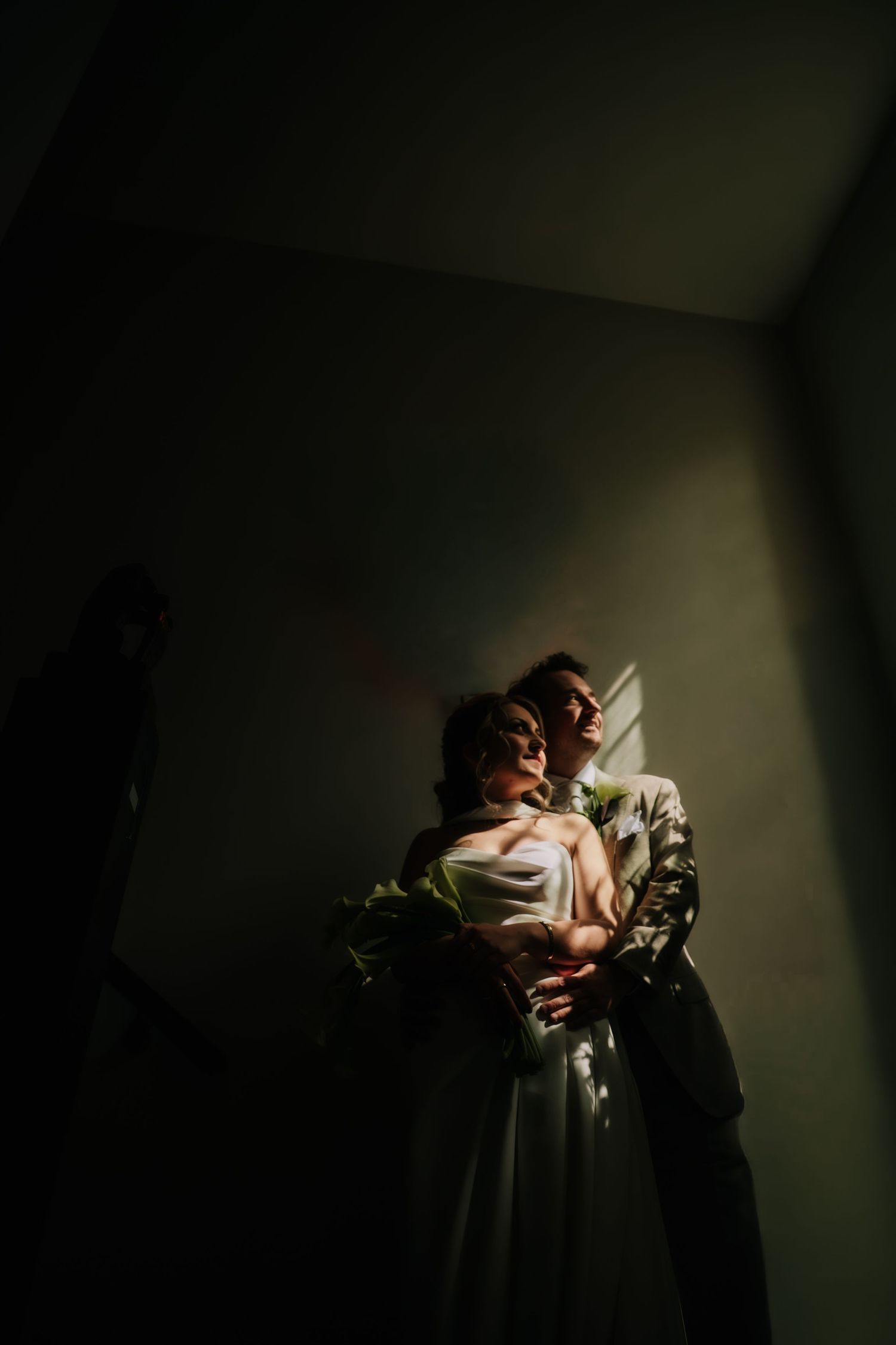 A bride and groom embrace in dramatic shadow and light near a corner wall, bride holding a green bouquet in wedding attire.