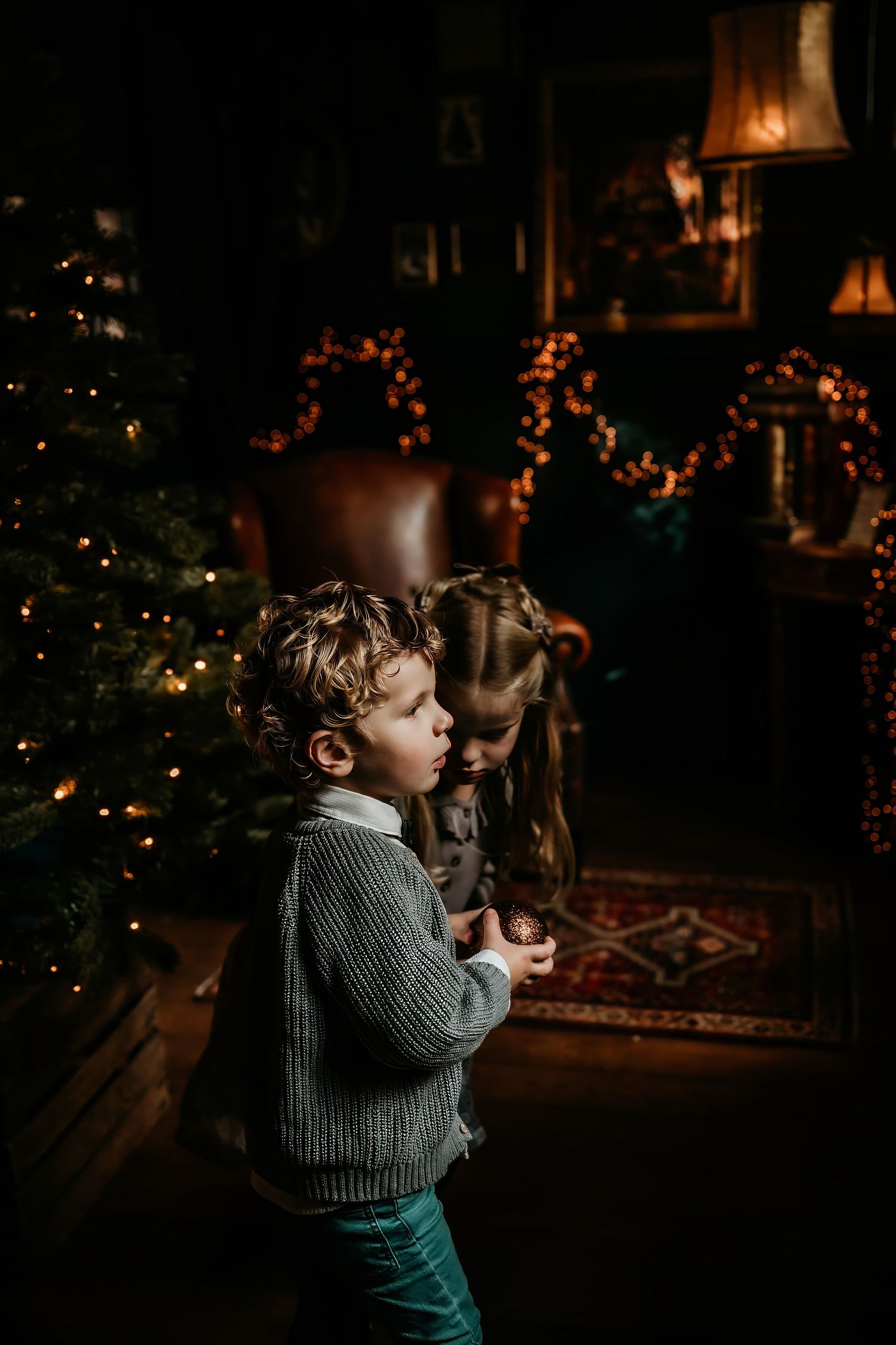 Child in a gray sweater gazes at twinkling Christmas lights in a cozy, dimly lit room with holiday decorations.