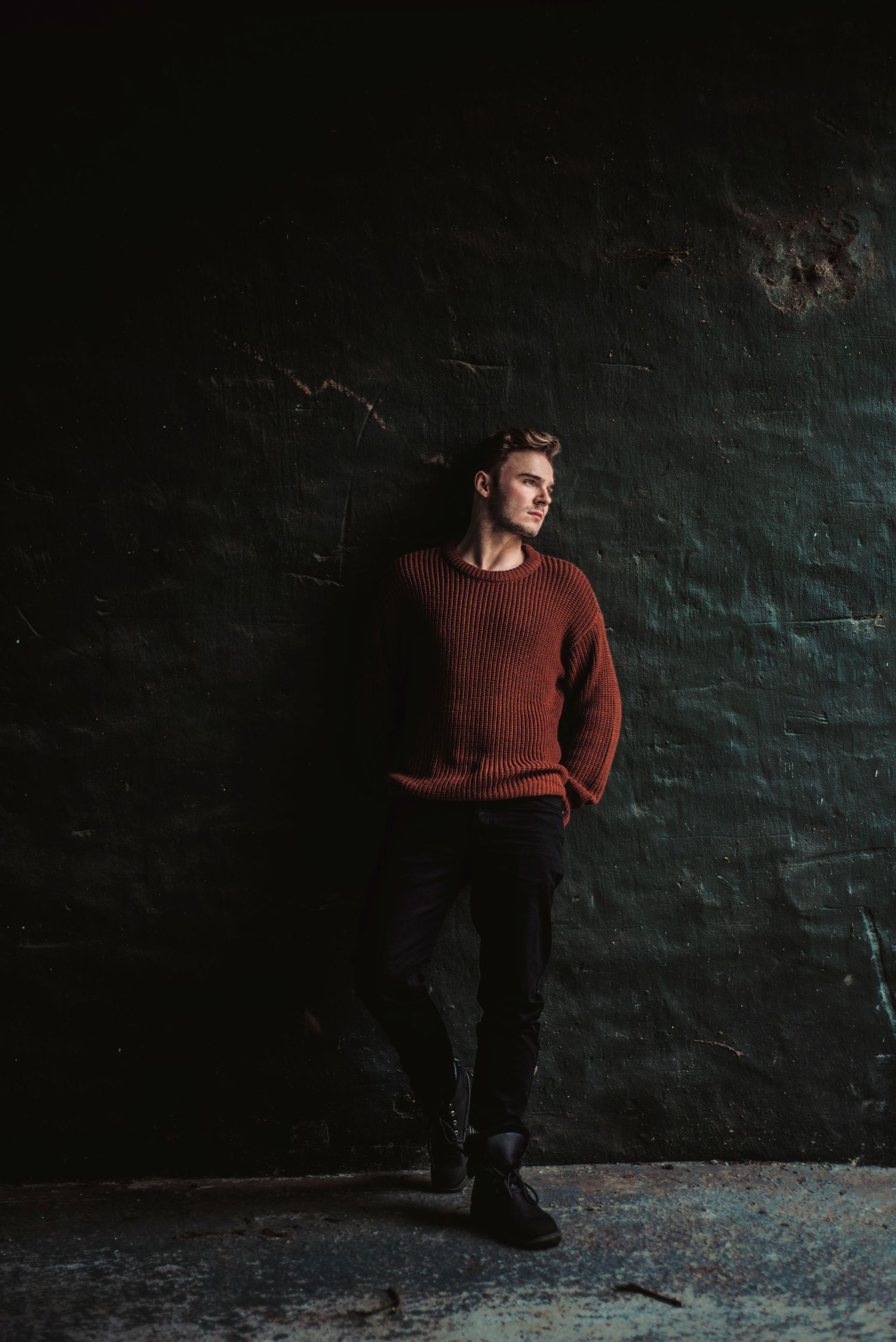 A person in a burgundy sweater poses against a dark textured wall in moody lighting.