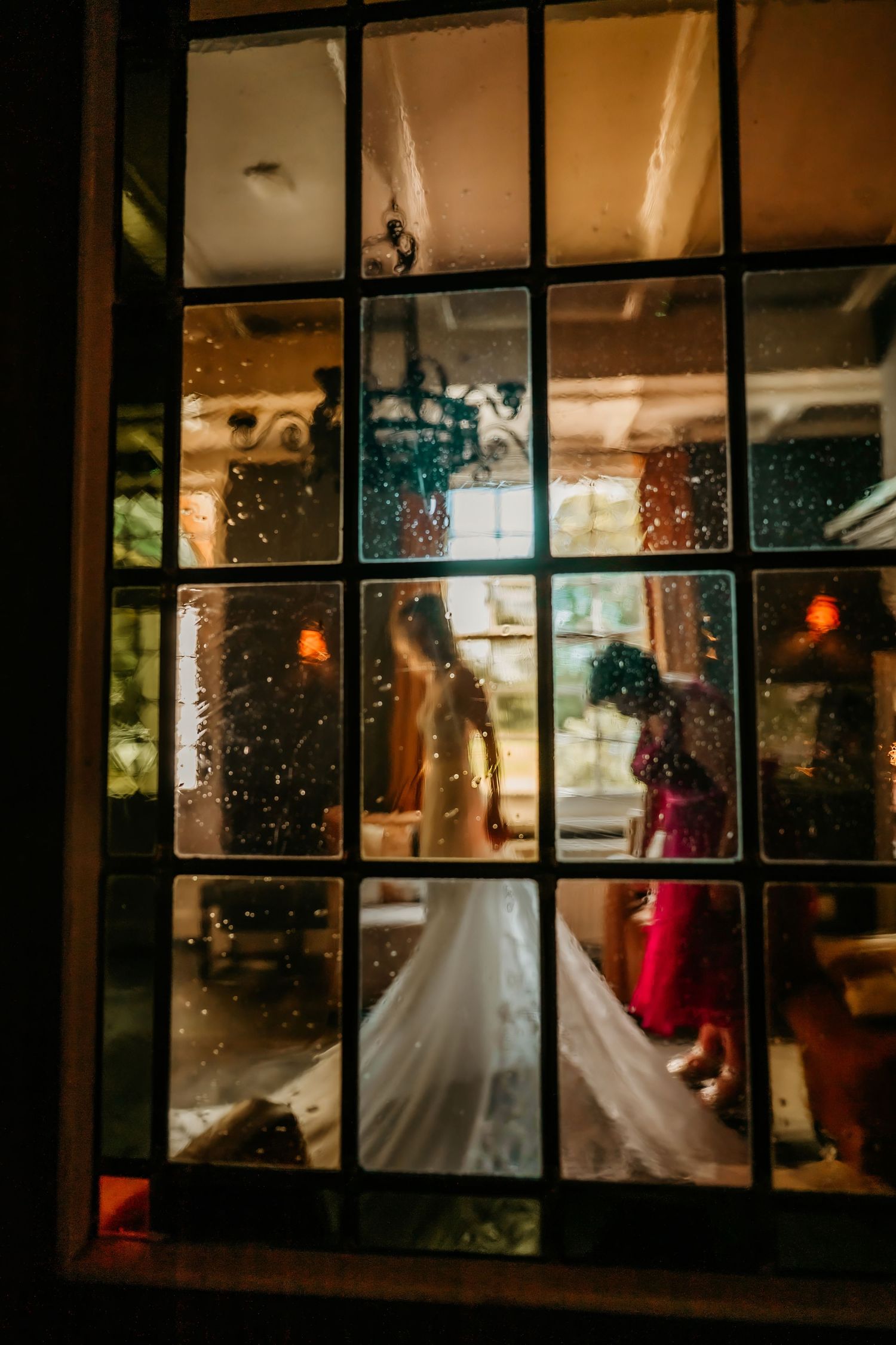 Wedding celebration viewed through rain-speckled window panes with warm interior lighting.