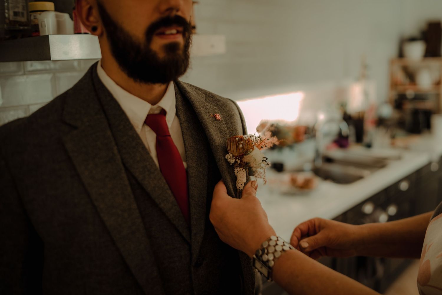Moody wedding detail shots showing boutonniere, suit details and hands in warm lighting.