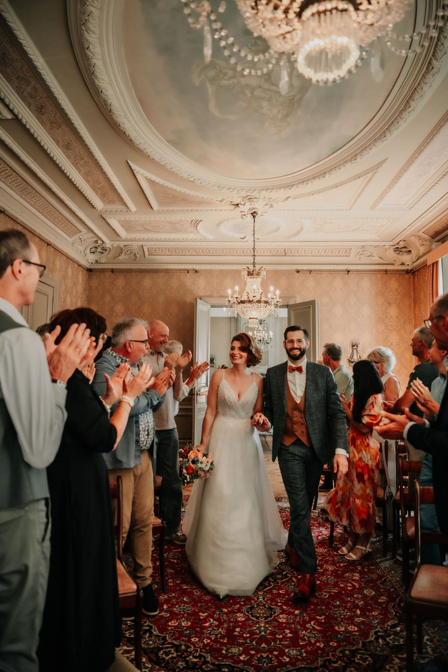 Wedding celebration in an ornate room with decorative ceiling and chandelier as guests applaud.