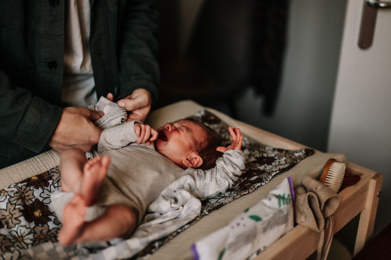 A newborn baby lies on a changing table wearing a light colored onesie.