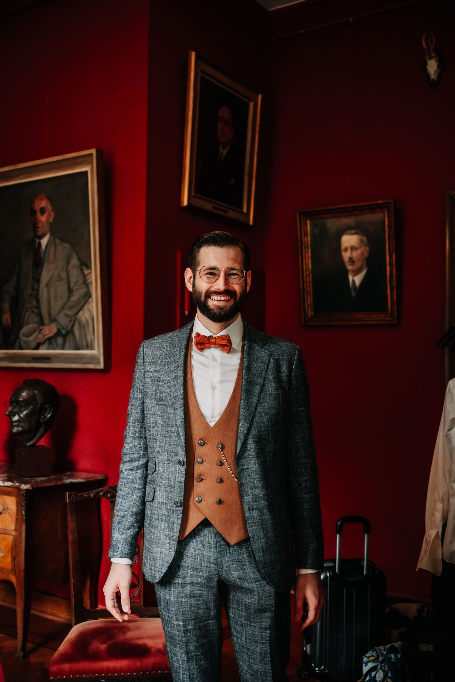 A stylish groom poses in a grey plaid suit with brown vest and red bow tie against a deep red wall with portraits.