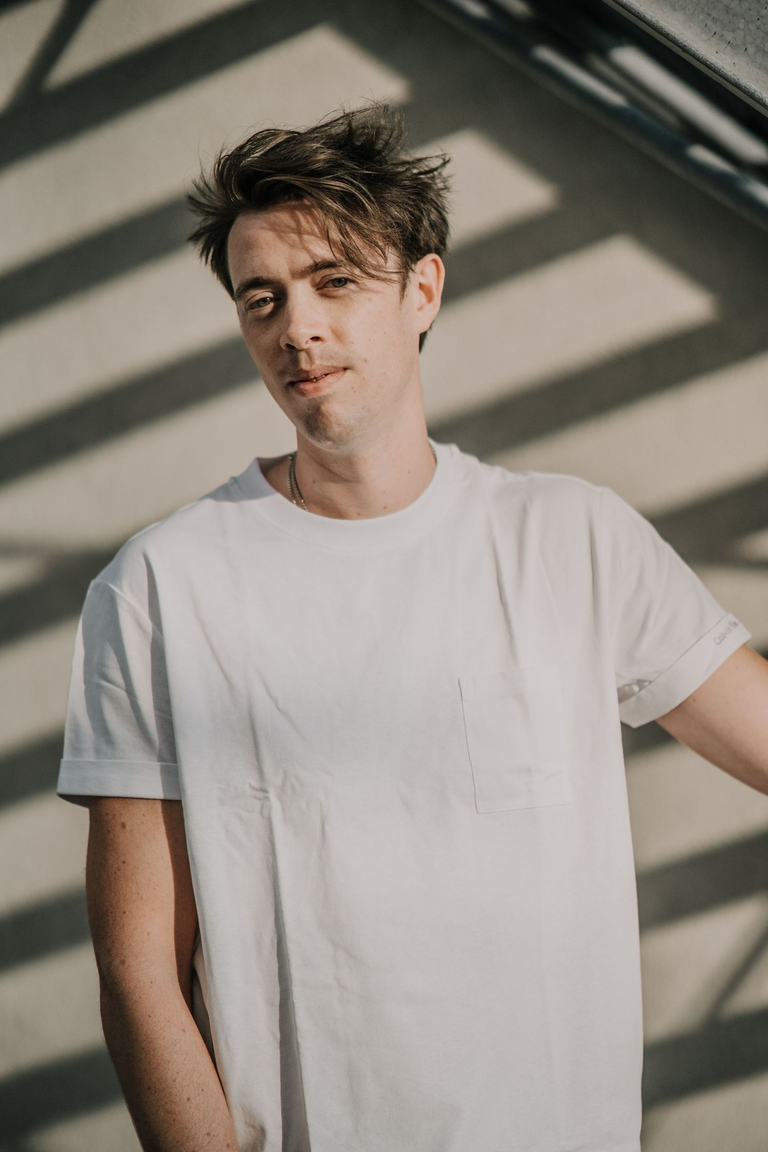 Person in white t-shirt poses against wall with dramatic shadow patterns creating artistic lighting effect.