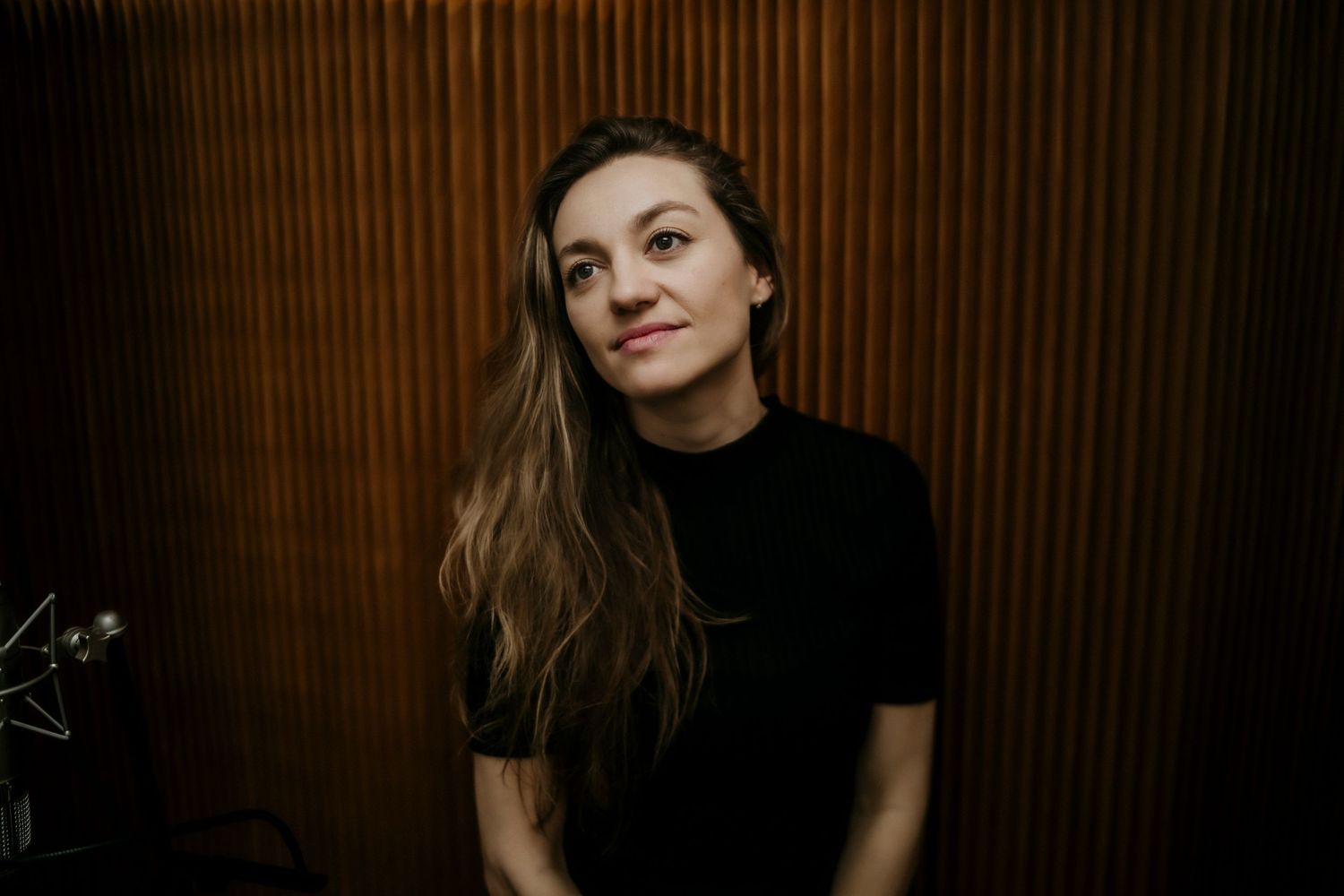 Person wearing a black top poses against a dark brown wooden wall in moody lighting.