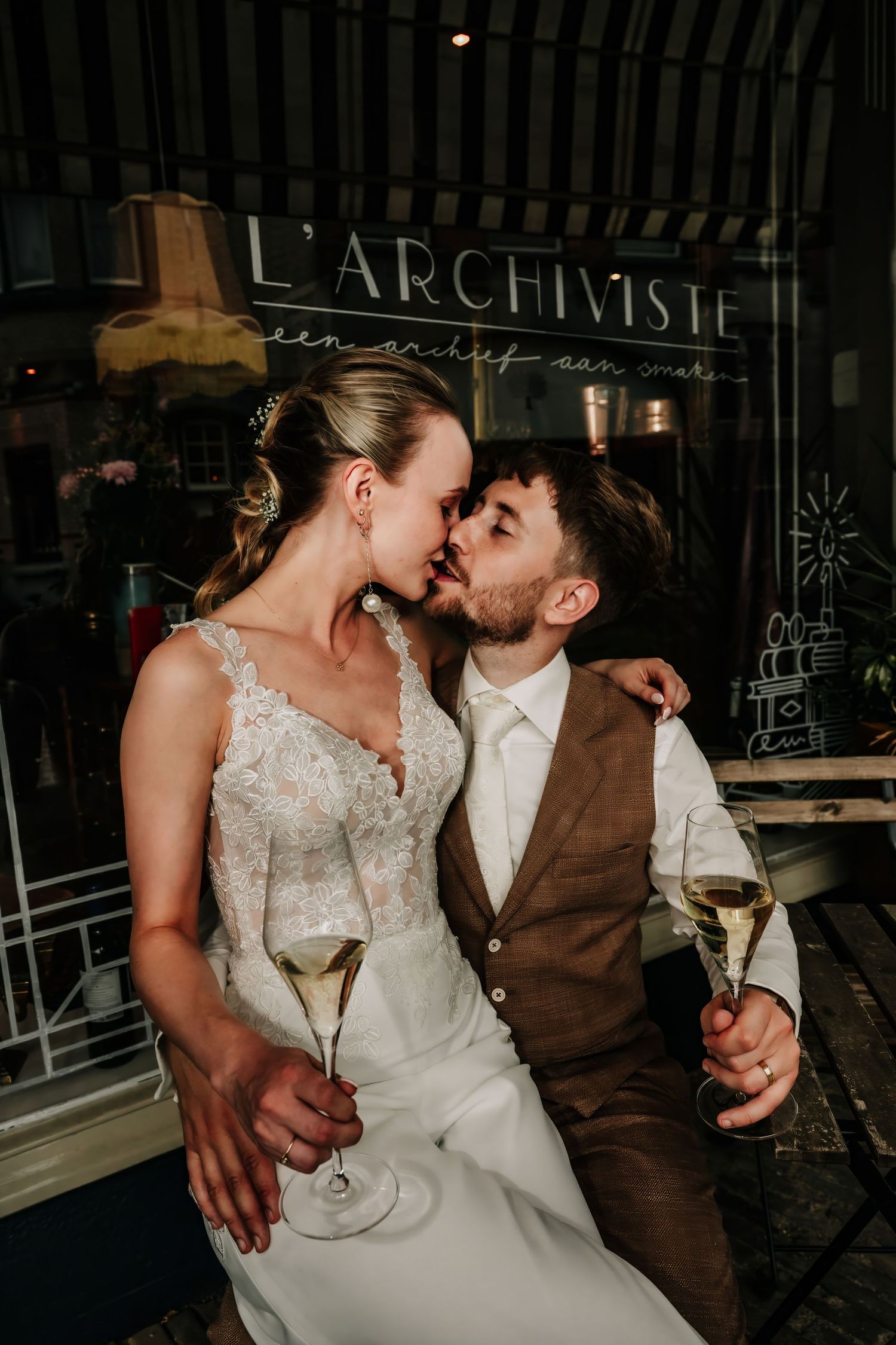Bride and groom kissing while holding champagne glasses outside L'Archiviste restaurant at their wedding reception.