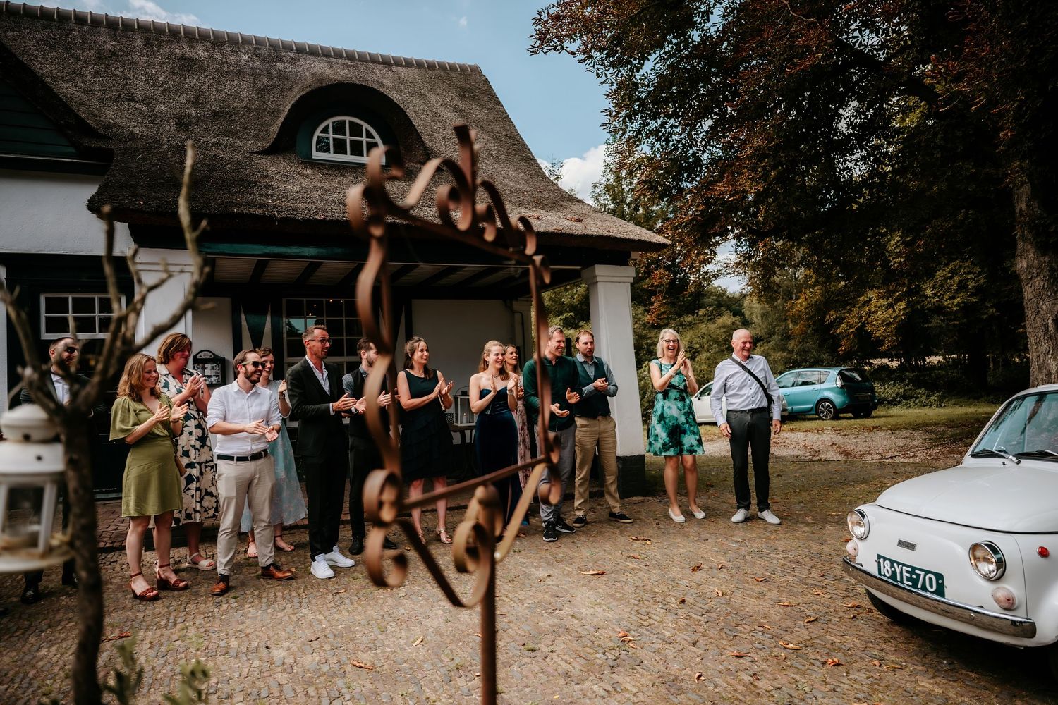 Joyful wedding guests celebrating outside a rustic thatched-roof cottage.