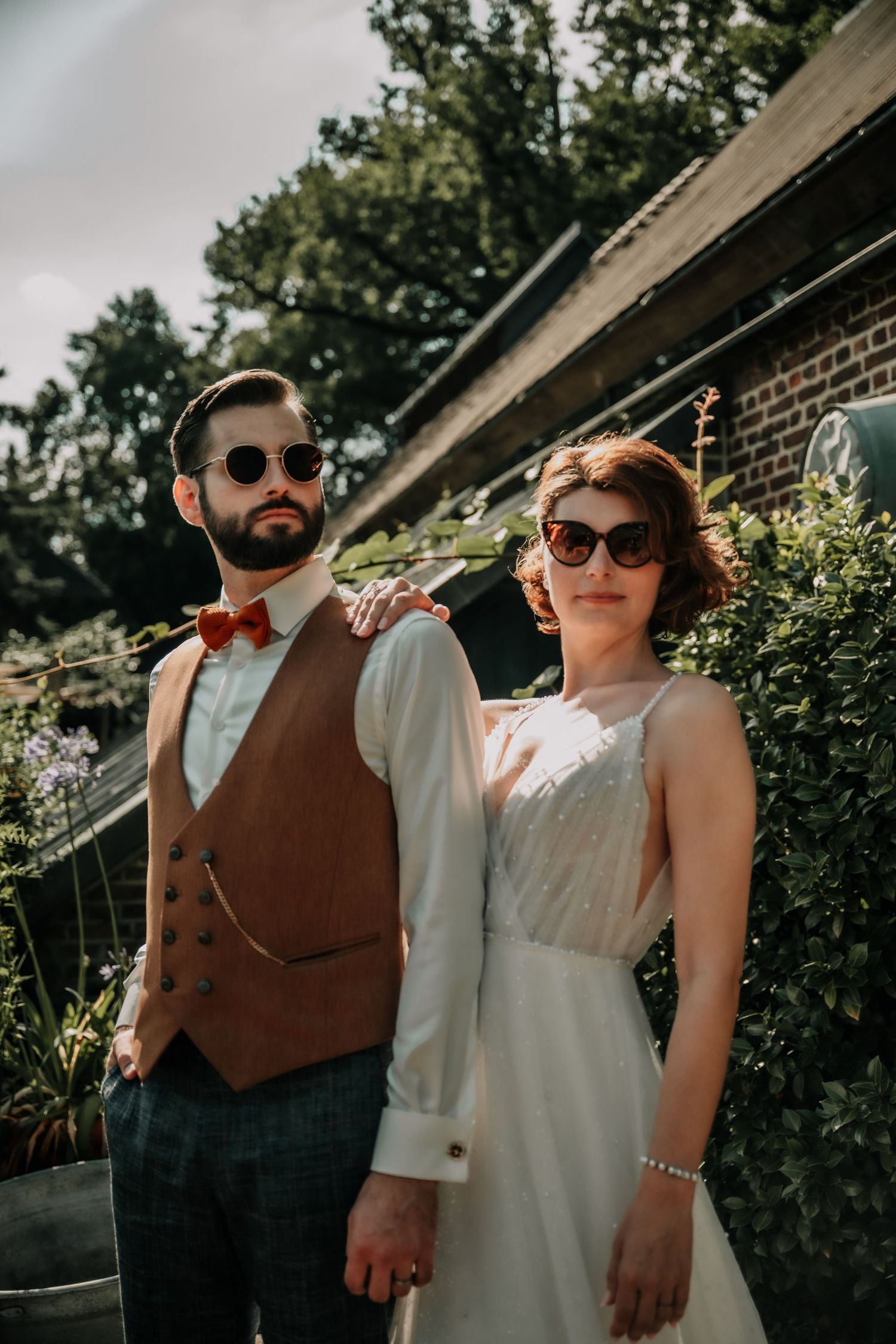 Stylish wedding couple poses outdoors wearing sunglasses against a brick wall backdrop.