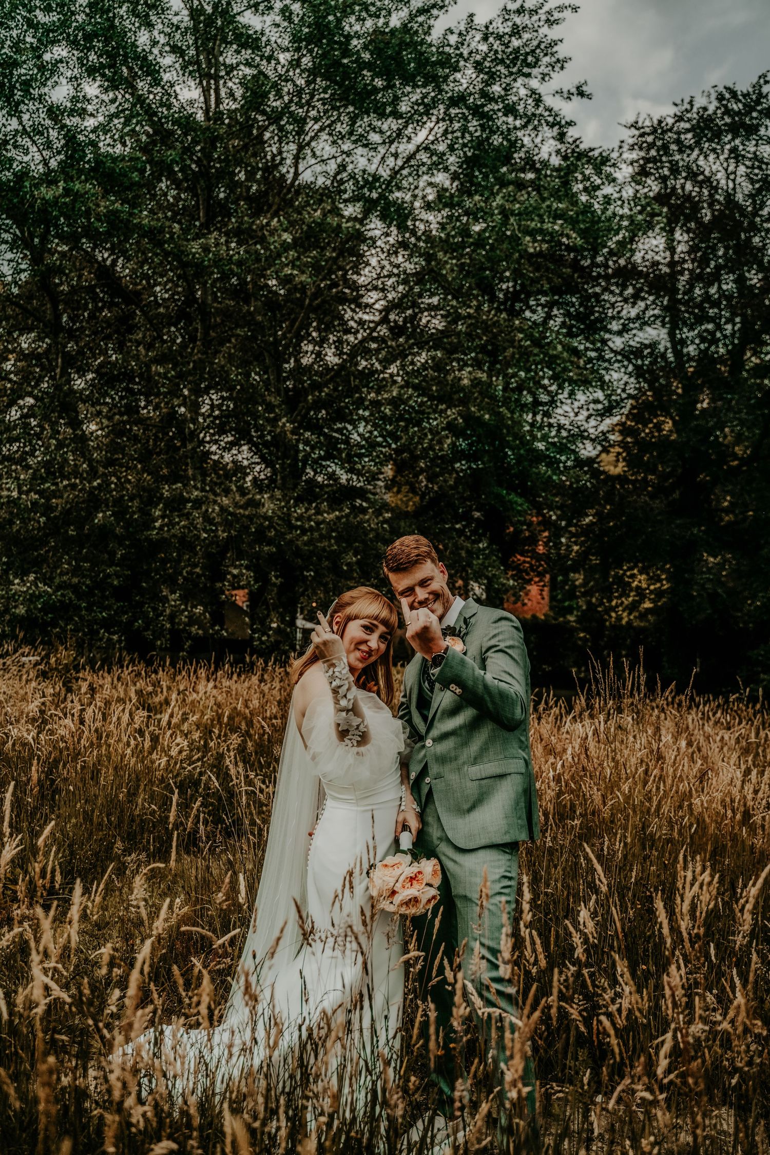 A wedding couple poses together in a golden field at sunset with trees in the background.
