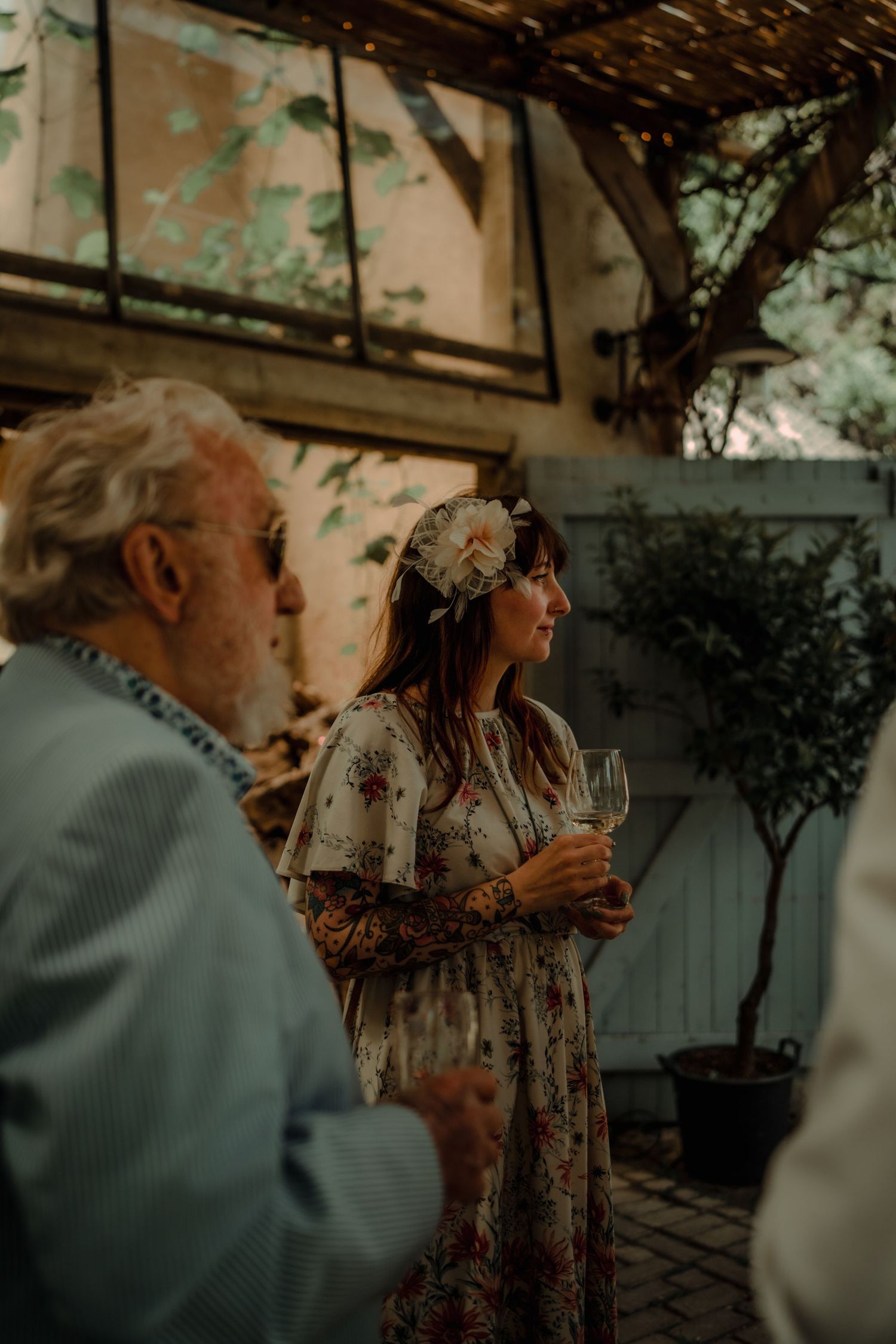 A gathering at an outdoor garden party with guests enjoying drinks under a vine-covered pergola.