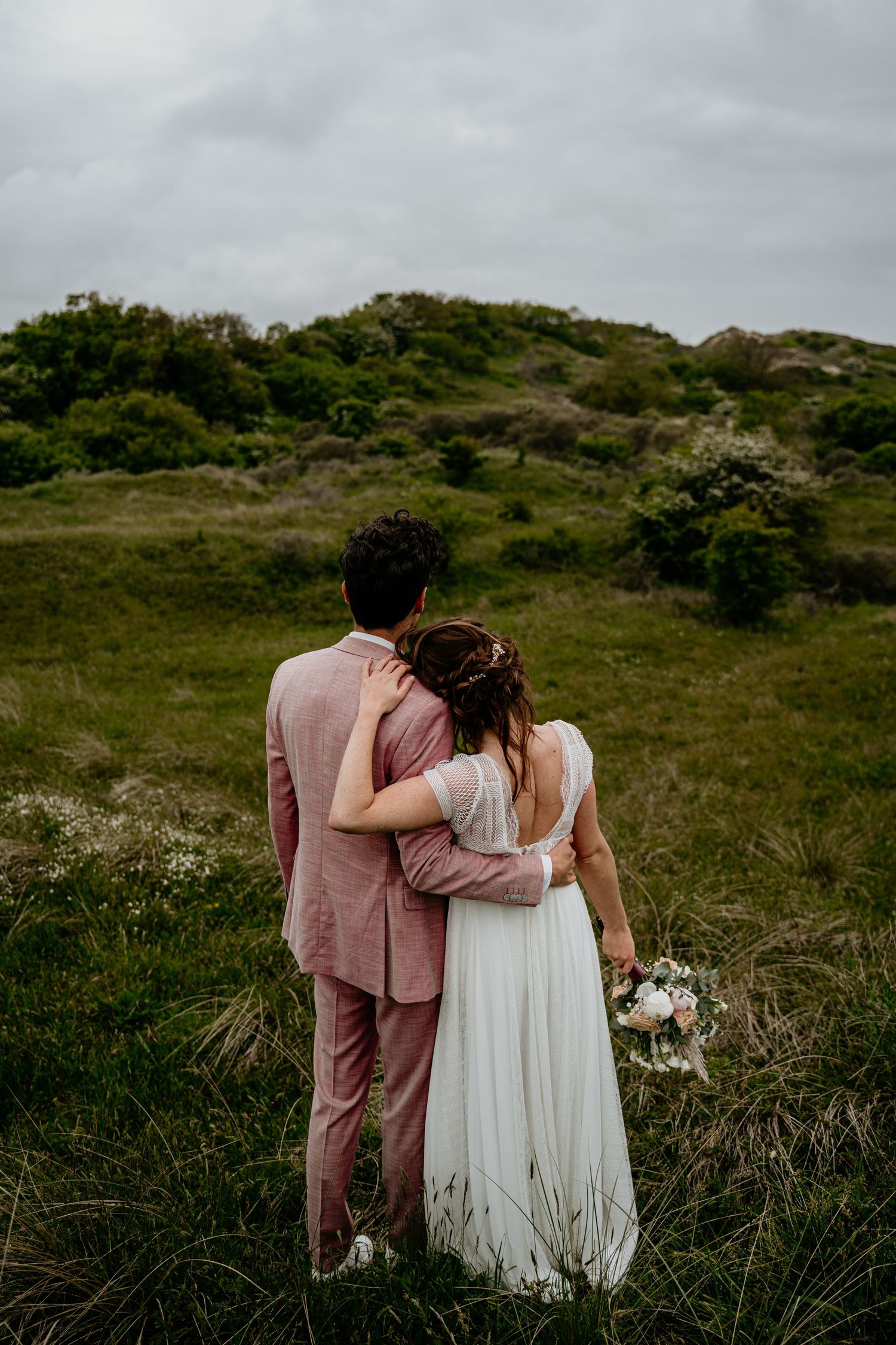 A couple in pink and white embrace while walking through a grassy coastal landscape.