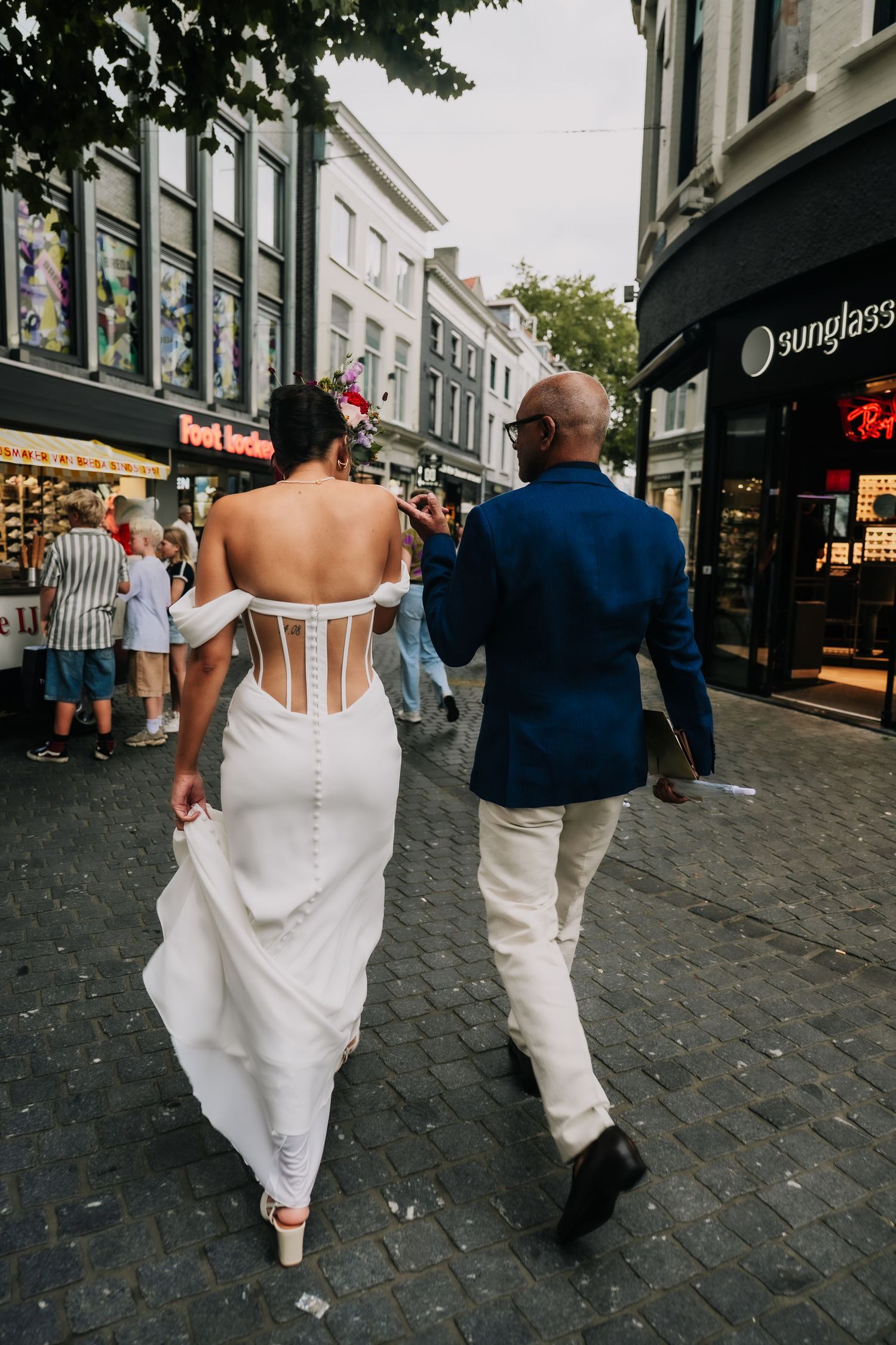 Bride in backless white gown walks with groom in navy suit through a European cobblestone shopping street.