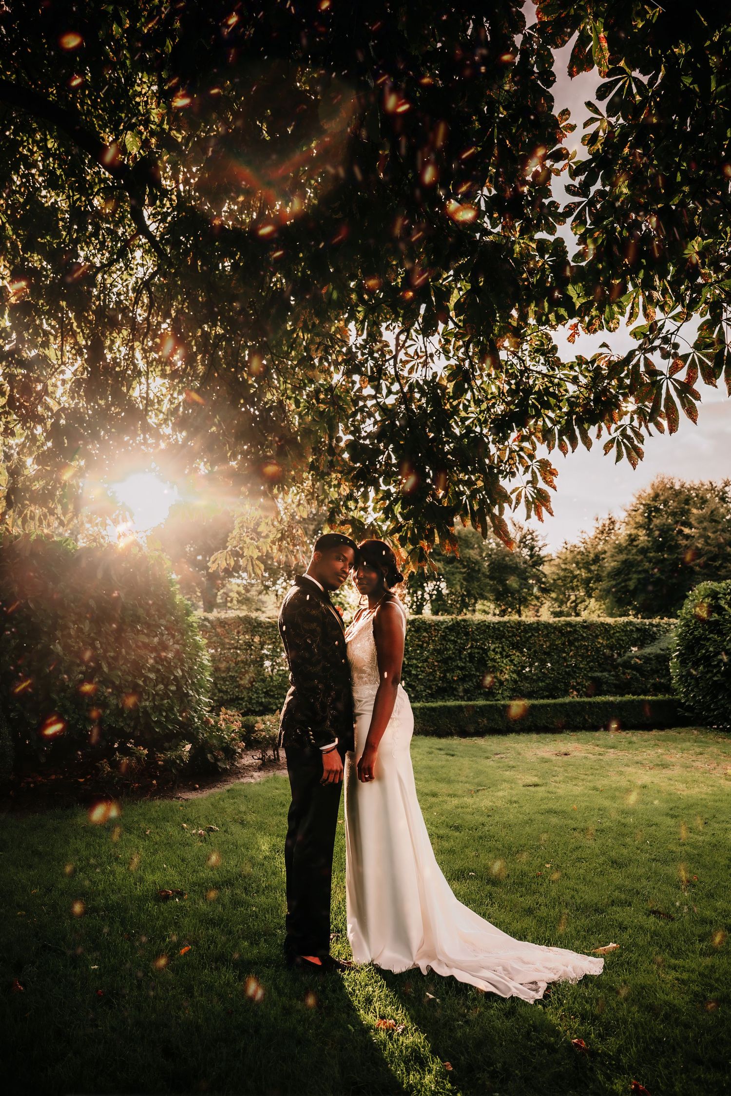 A romantic wedding couple stands in a sunlit garden with golden light filtering through autumn leaves at sunset.