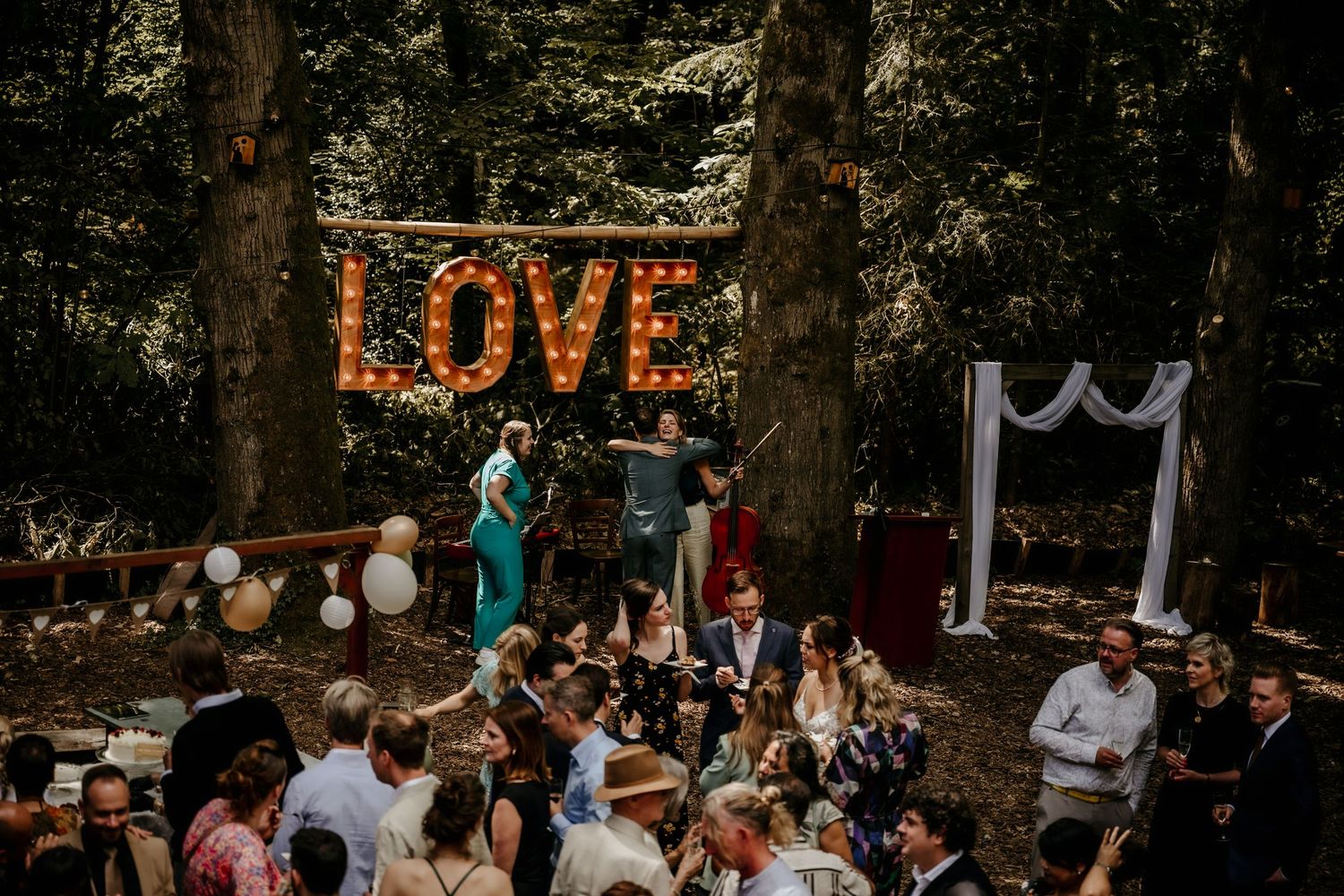 String lights illuminate an outdoor wedding reception celebration with guests mingling under a forest canopy at dusk.