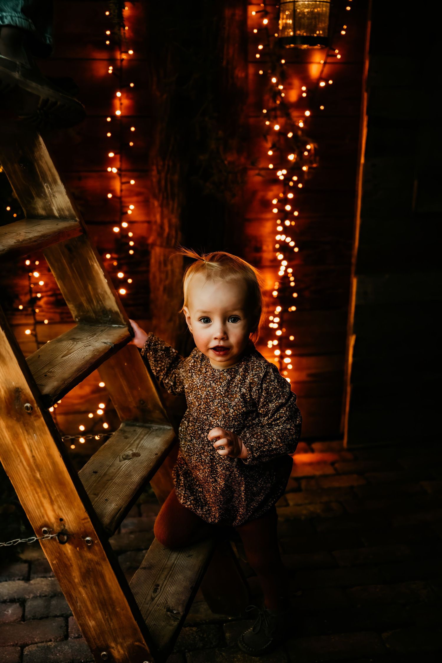 A child in a sparkly dress sits on wooden stairs with glowing string lights creating a magical holiday atmosphere.
