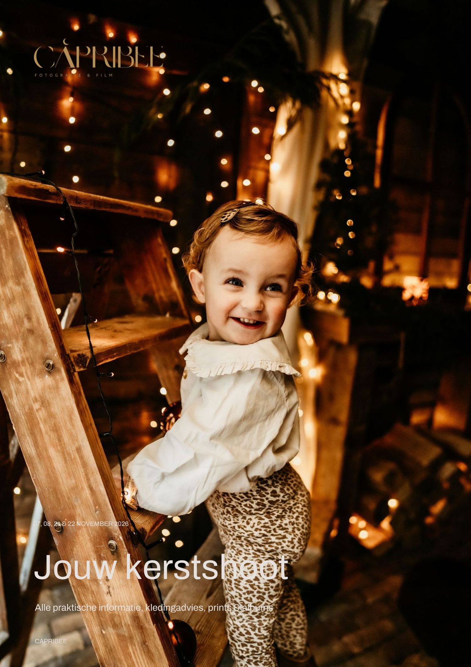 Smiling toddler in a white blouse and leopard-print pants stands by a wooden ladder with warm fairy lights at a cozy Christmas photoshoot.