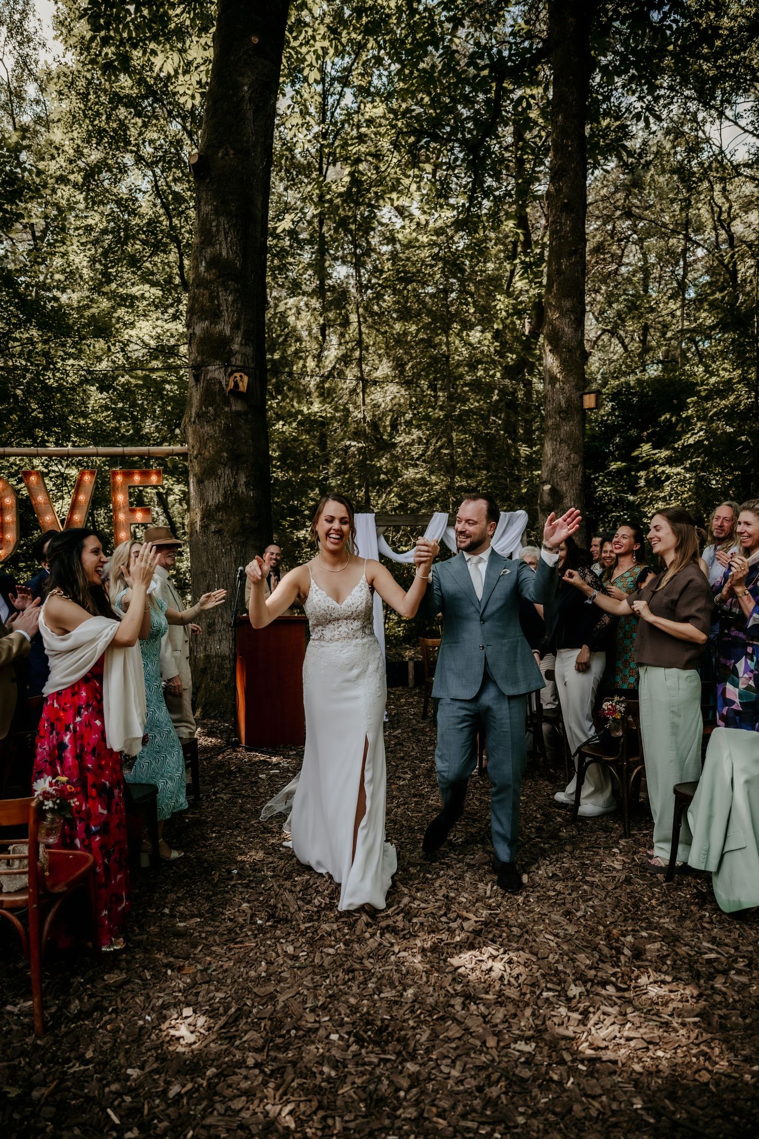 Wedding party celebrates with joyful dance moves in a wooded outdoor setting during golden hour.