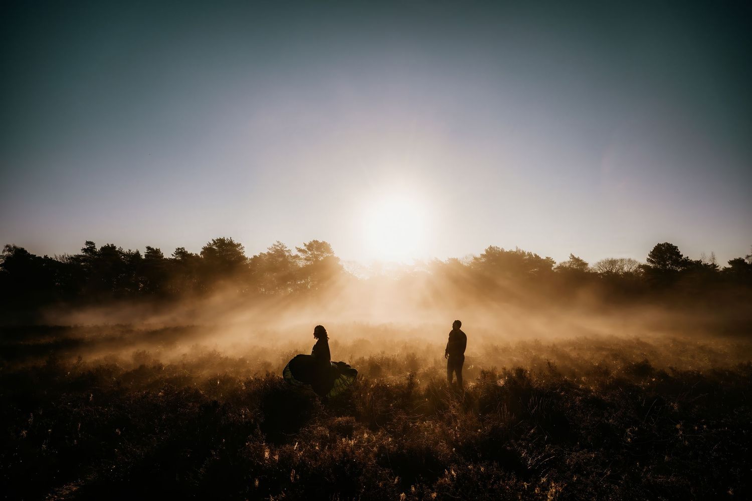 Silhouettes of figures in foggy landscape during dramatic sunrise with sun peeking through haze.