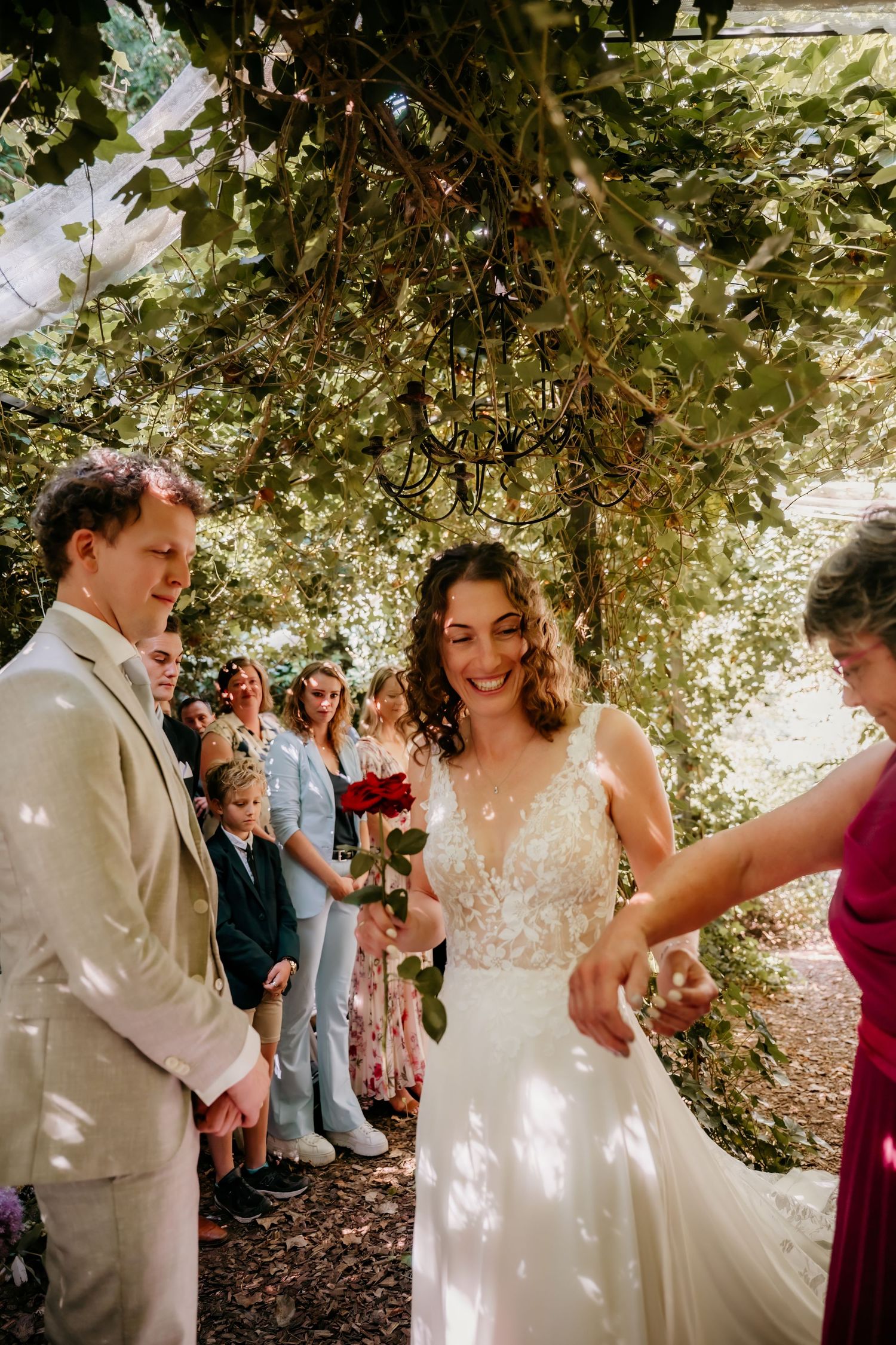 A wedding ceremony takes place under a leafy garden canopy with guests looking on.