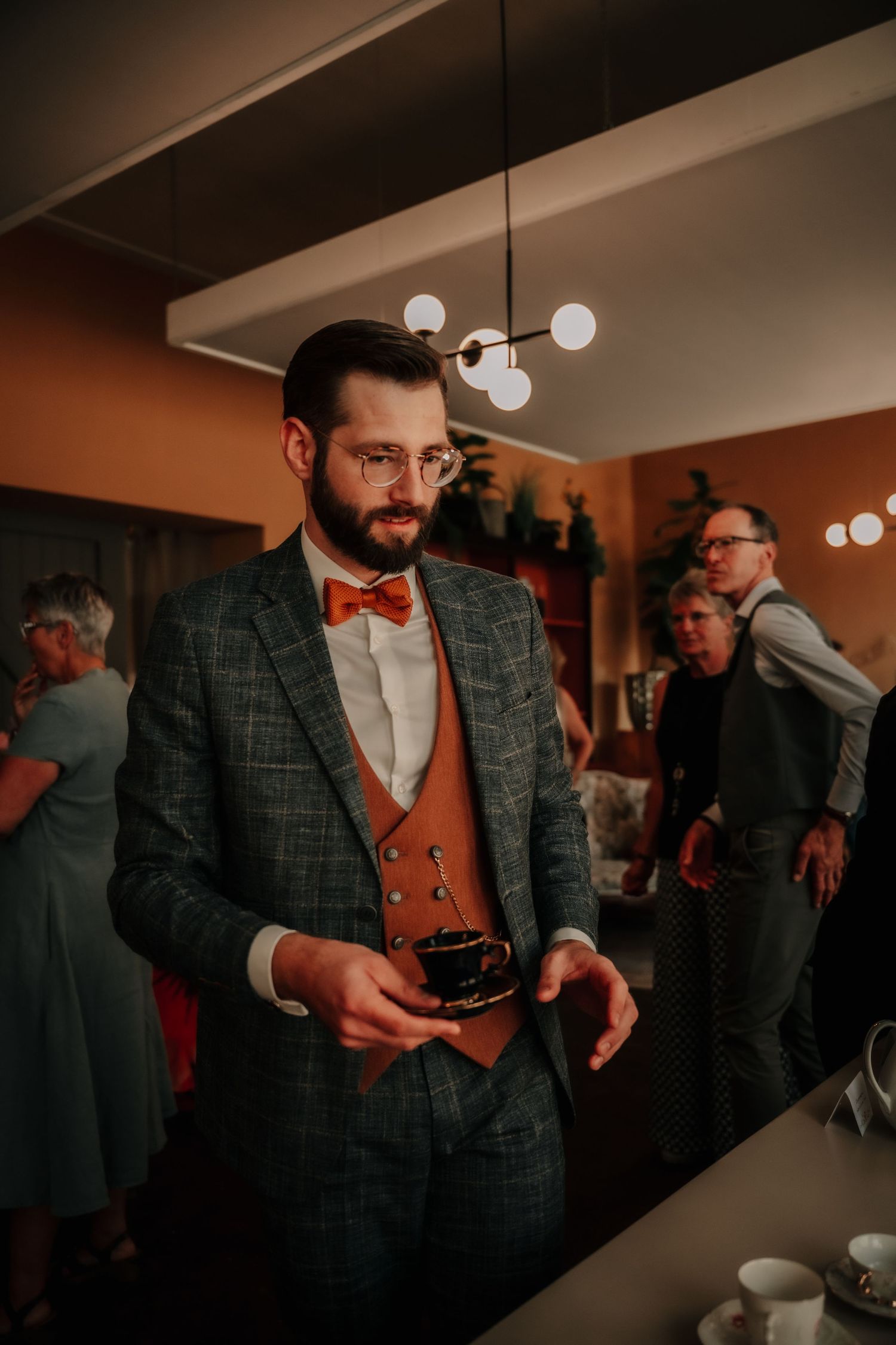 A stylishly dressed person in a plaid suit and red bowtie holds a camera at an indoor event.