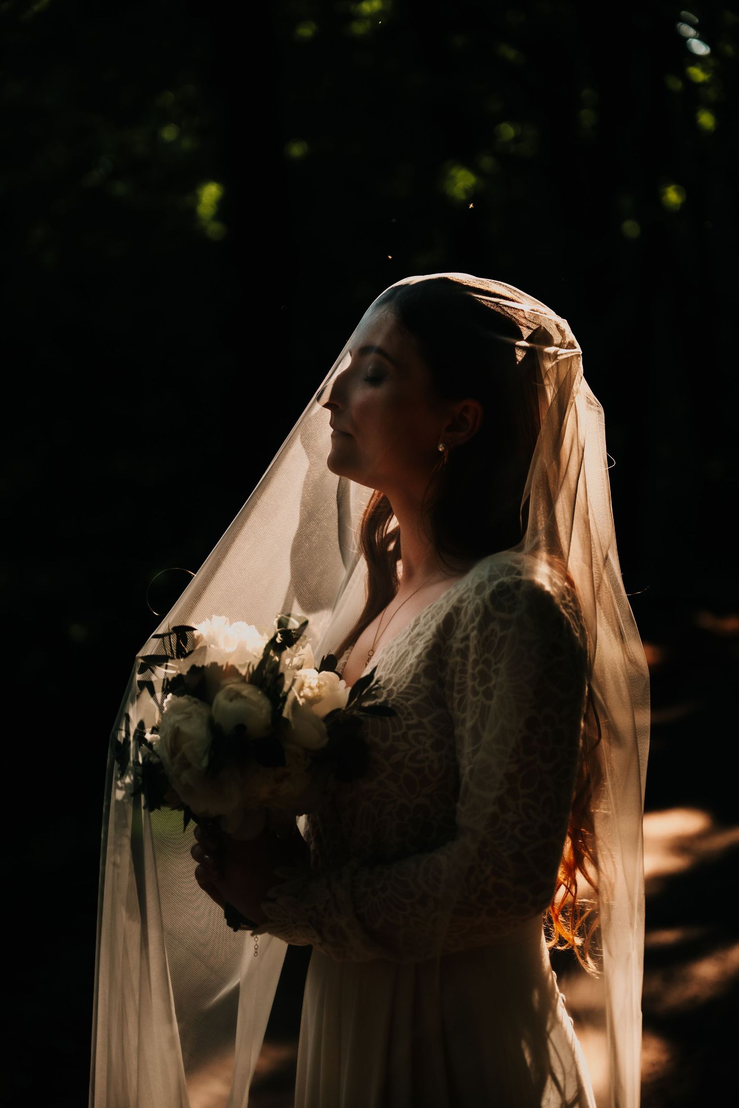 Bride in lace dress and veil holds bouquet in dramatic sunlit portrait.