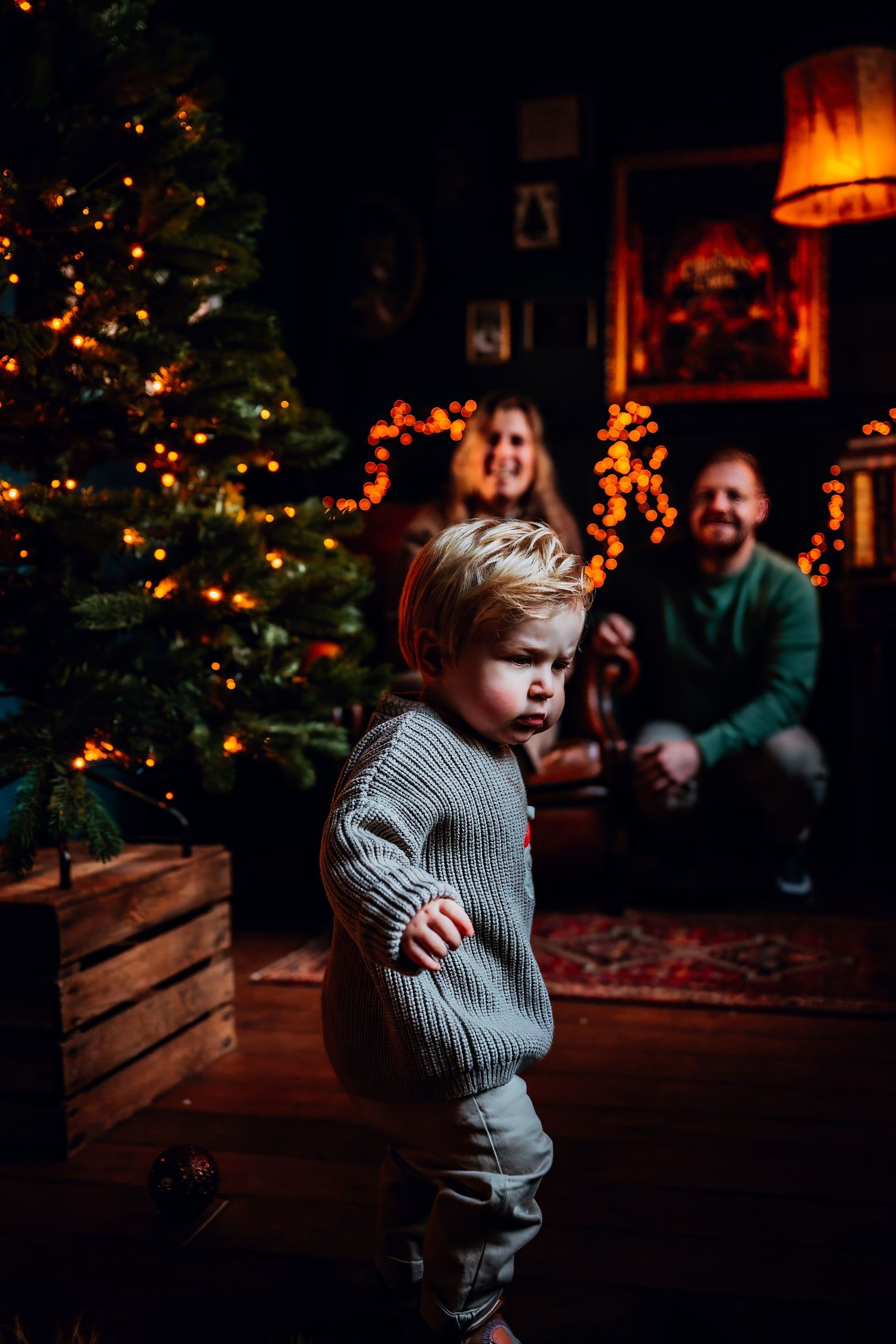 A toddler in a gray sweater stands in a dimly lit room with a Christmas tree glowing in the background.