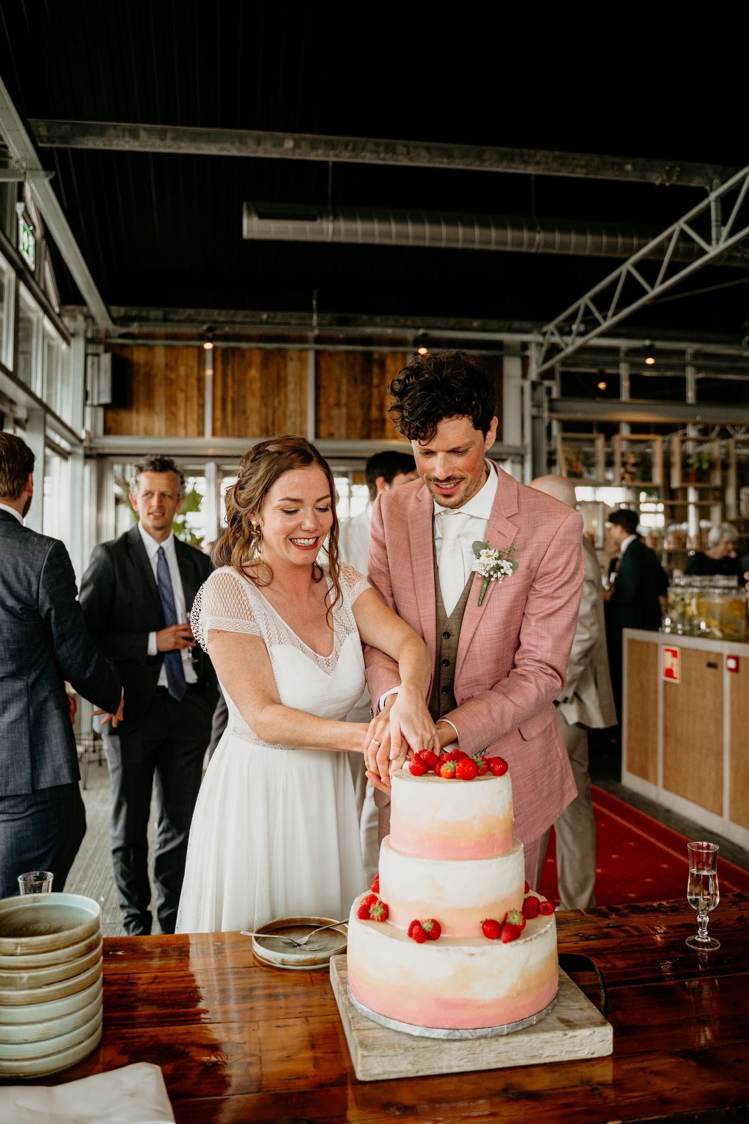 A wedding couple cuts into their elegant white and coral-colored wedding cake at a rustic reception venue.