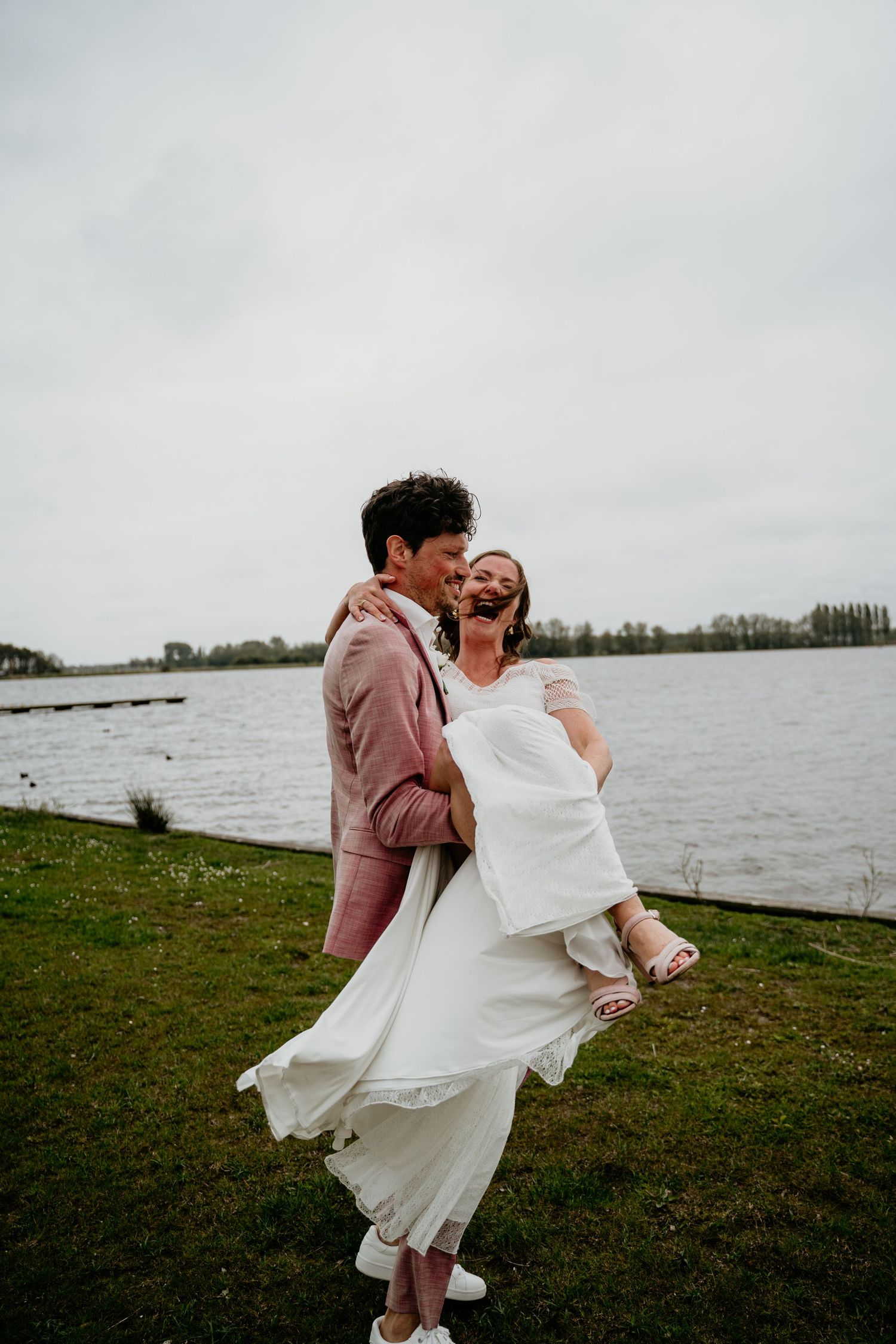 A romantic wedding couple embraces on a grassy shore beside a serene lake on an overcast day.