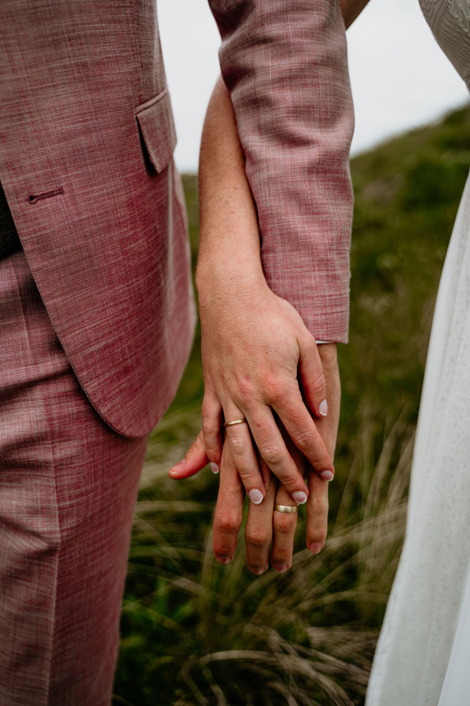 Close-up of hands wearing wedding rings against white dress and mauve suit fabric.