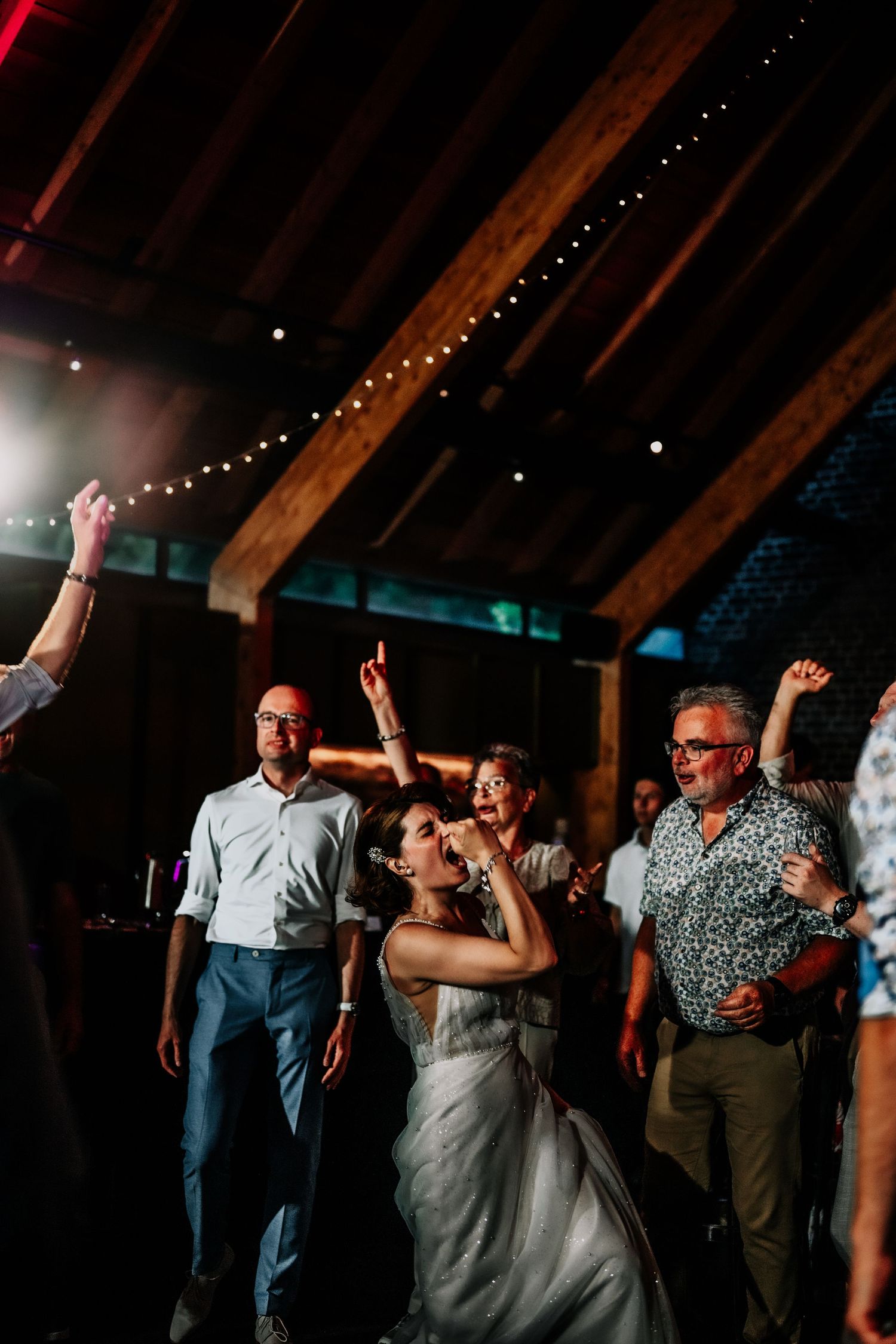 Wedding guests dancing and celebrating under string lights in a dimly lit barn venue.