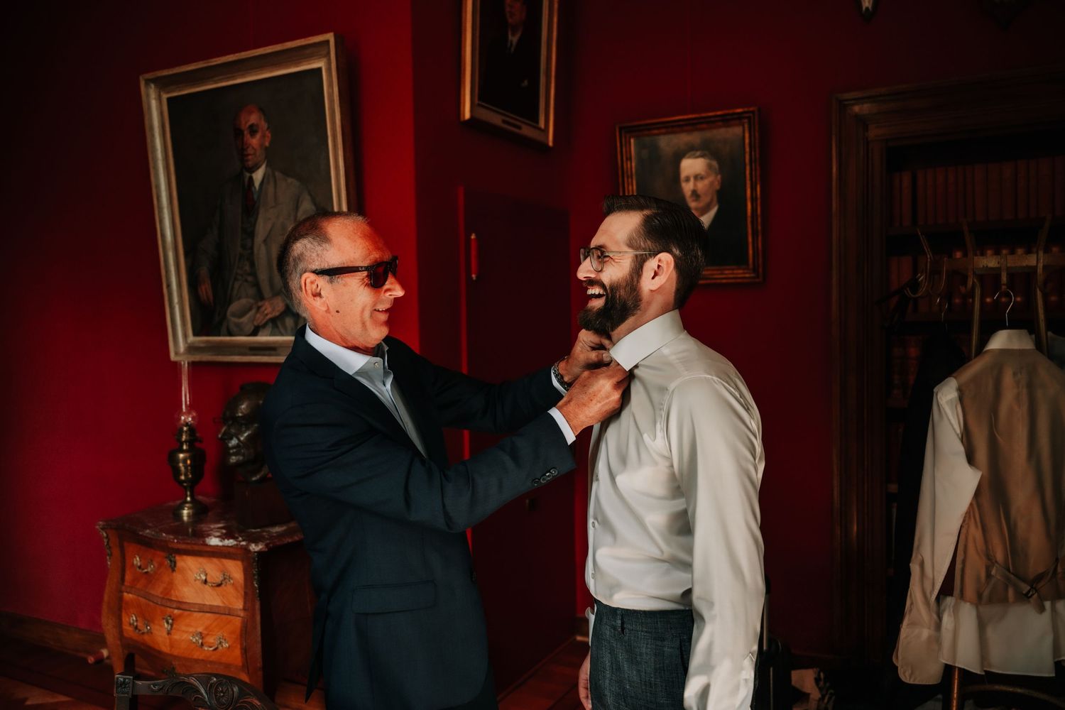 Two people in formal attire chat and laugh in front of a deep red wall with framed artwork.