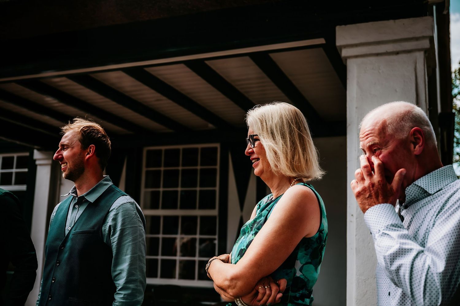 Three people stand on a covered porch in casual attire during a daytime gathering.