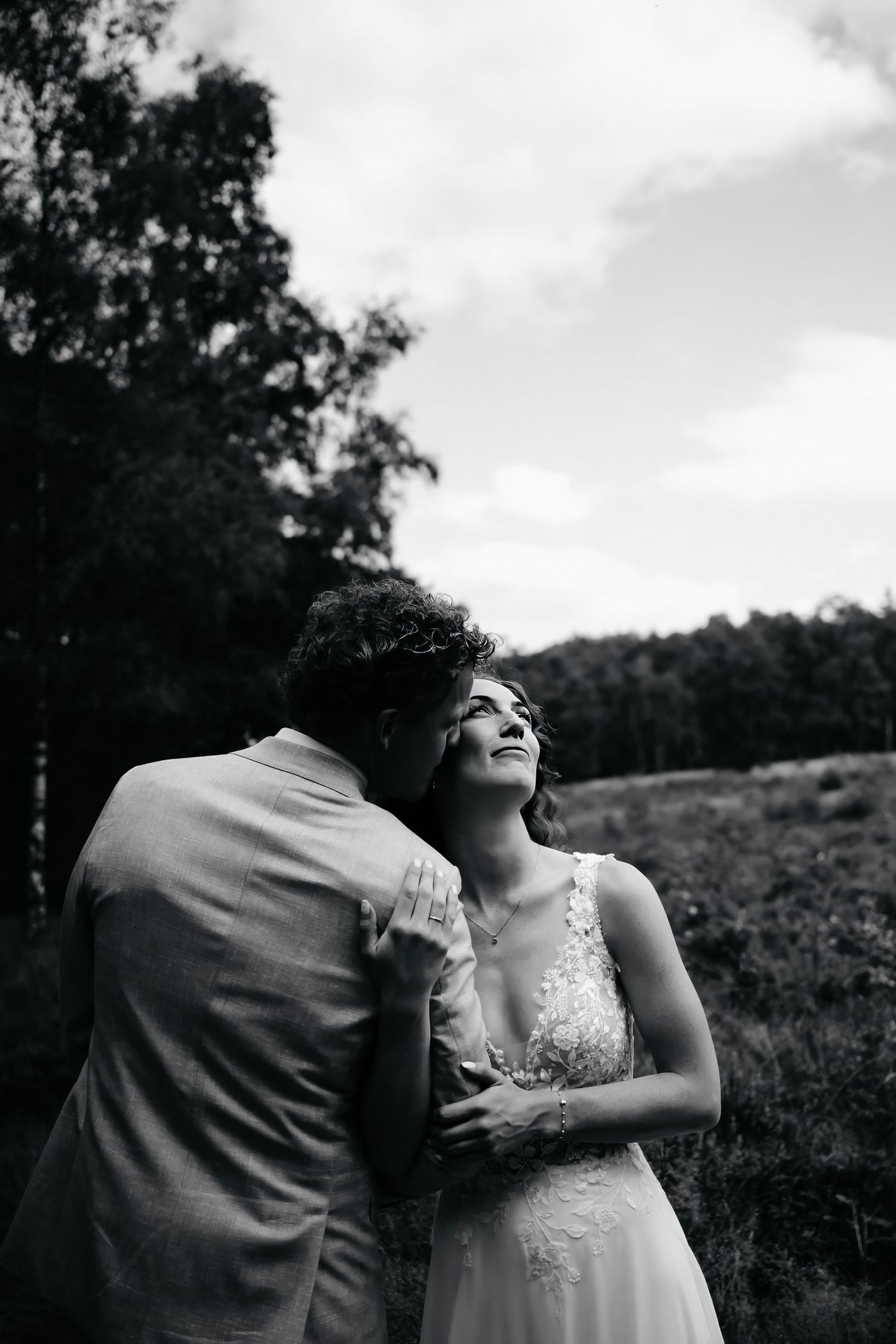 Intimate black and white wedding moment captured in outdoor field with cloudy sky backdrop.
