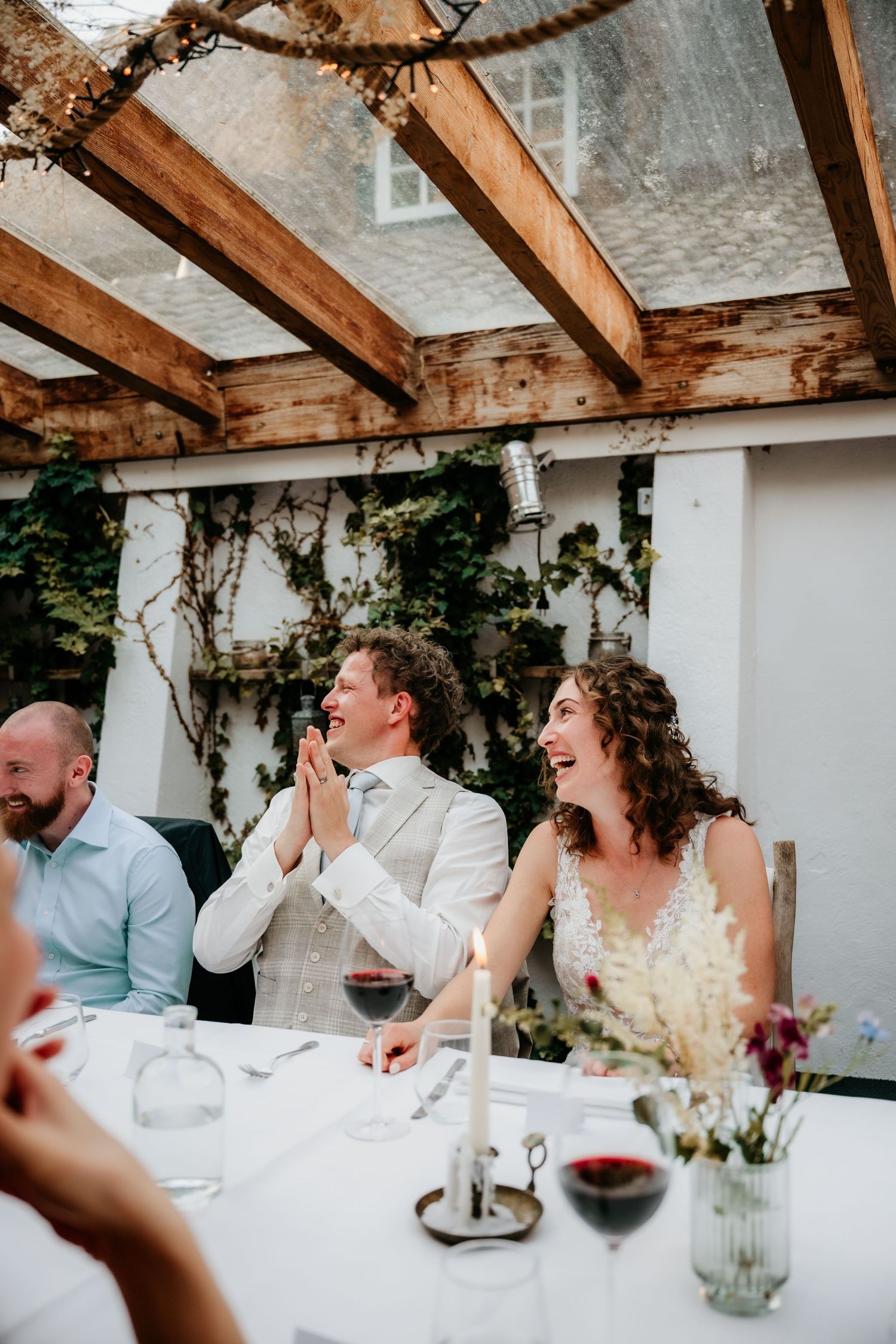 Guests sharing joyful laughter during a wedding reception under a rustic wooden pergola.