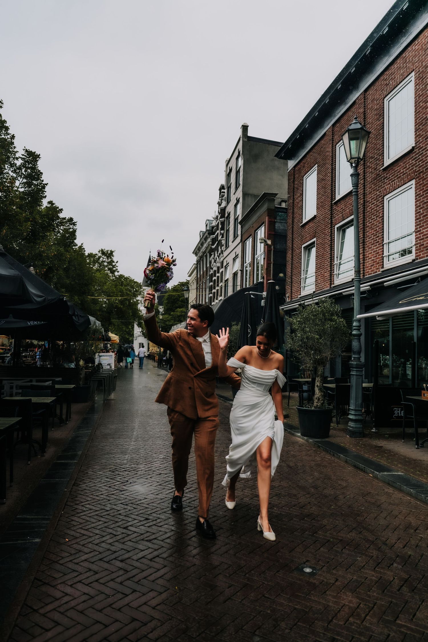 Bride and groom walking joyfully on a rainy cobblestone street, groom in brown suit holding colorful bouquet.