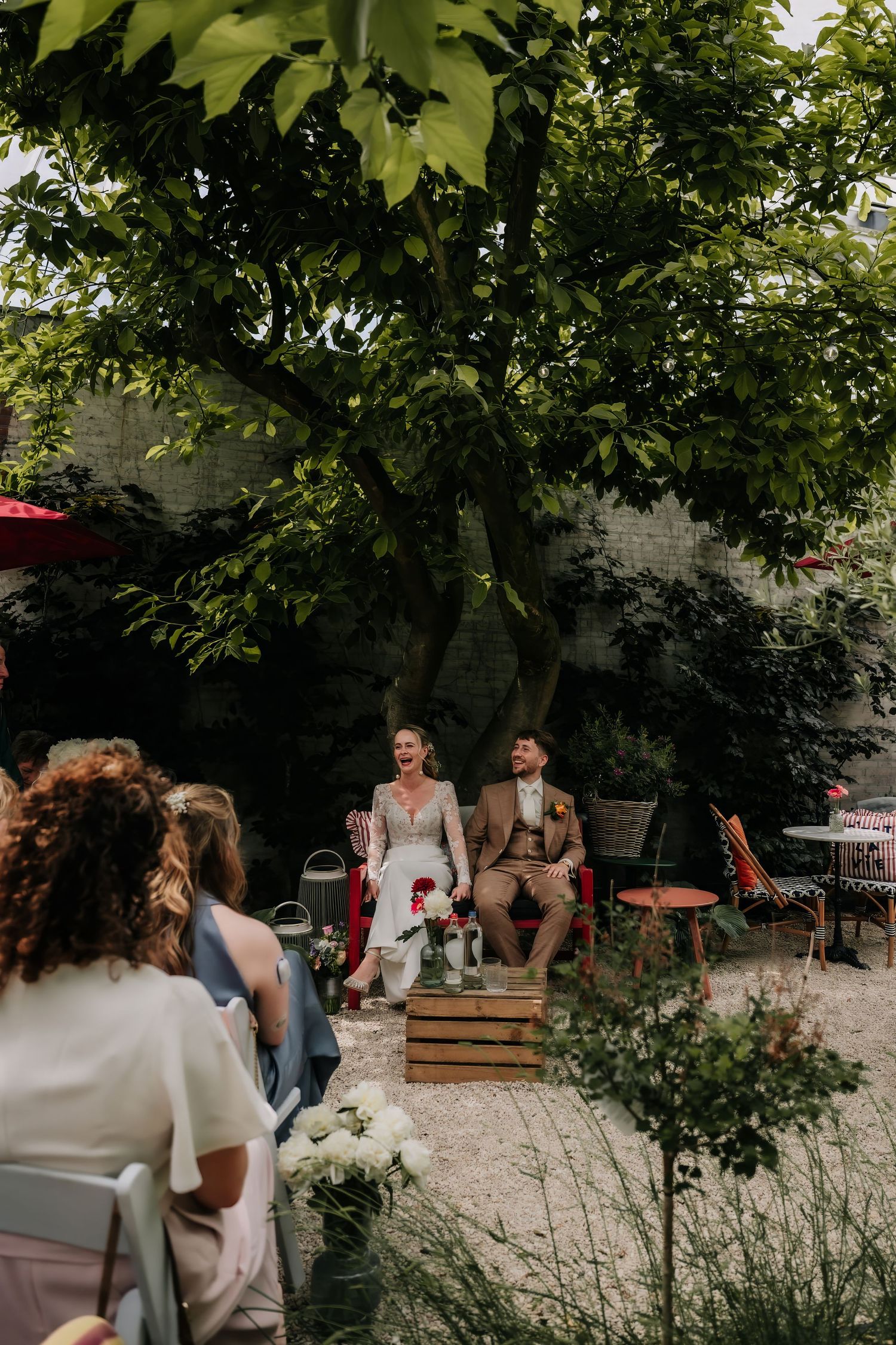 Couple laughing during outdoor garden wedding ceremony seated on wooden crates beneath lush green trees with guests watching.