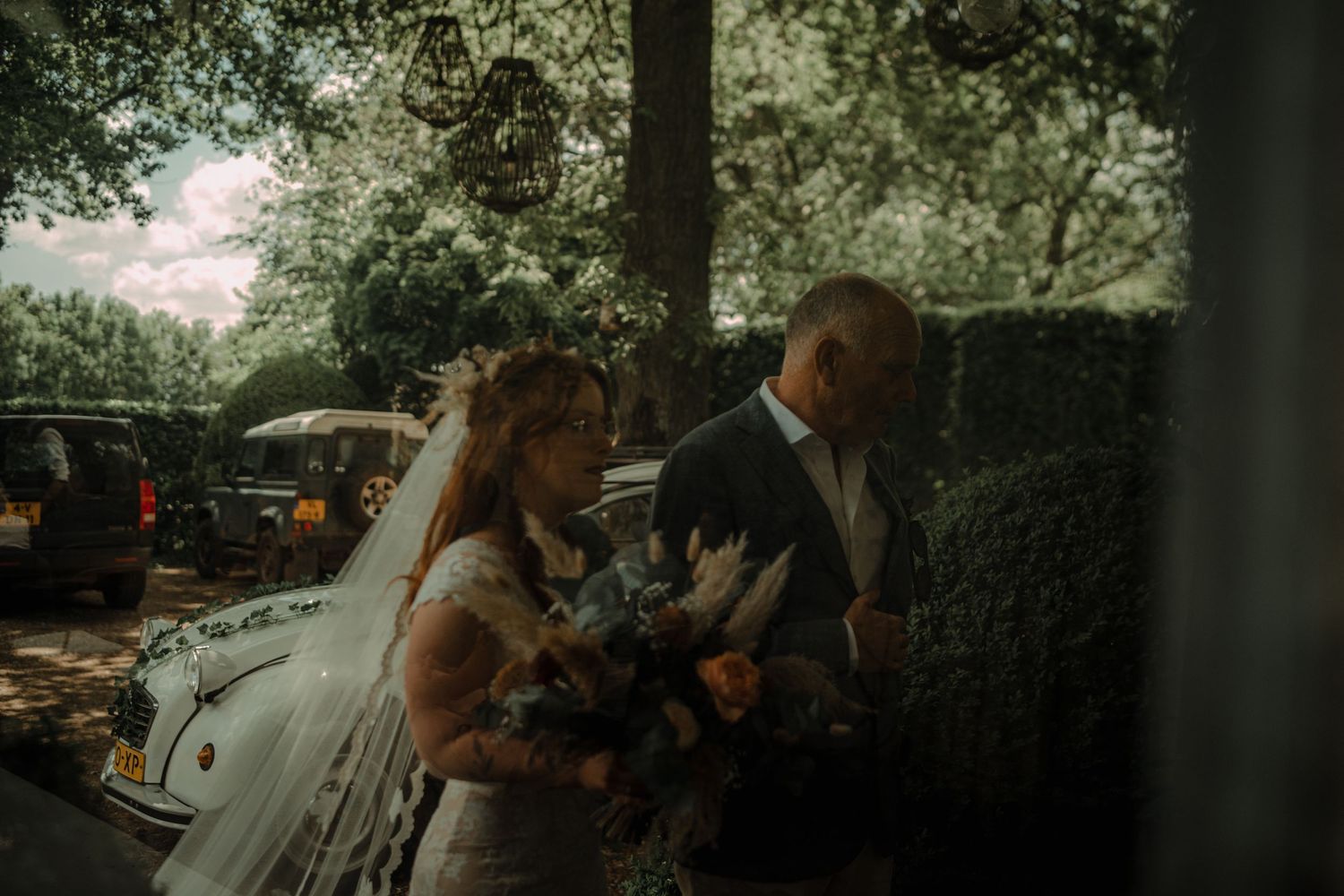 A wedding celebration scene in a garden setting with floral arrangements and vintage cars.