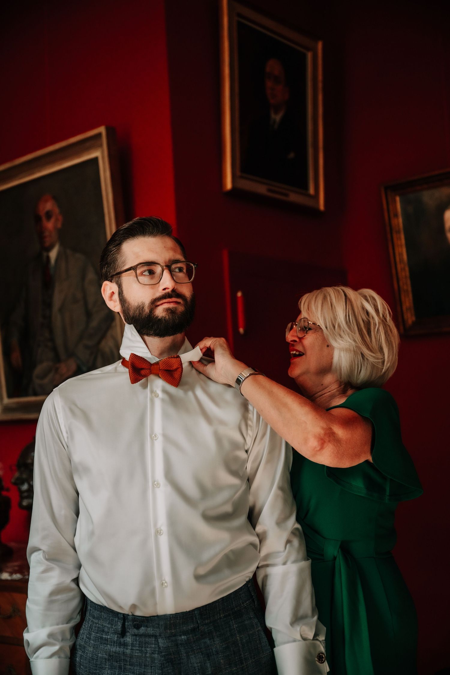Wedding preparation scene with a red bow tie adjustment in an elegant room with portrait paintings.